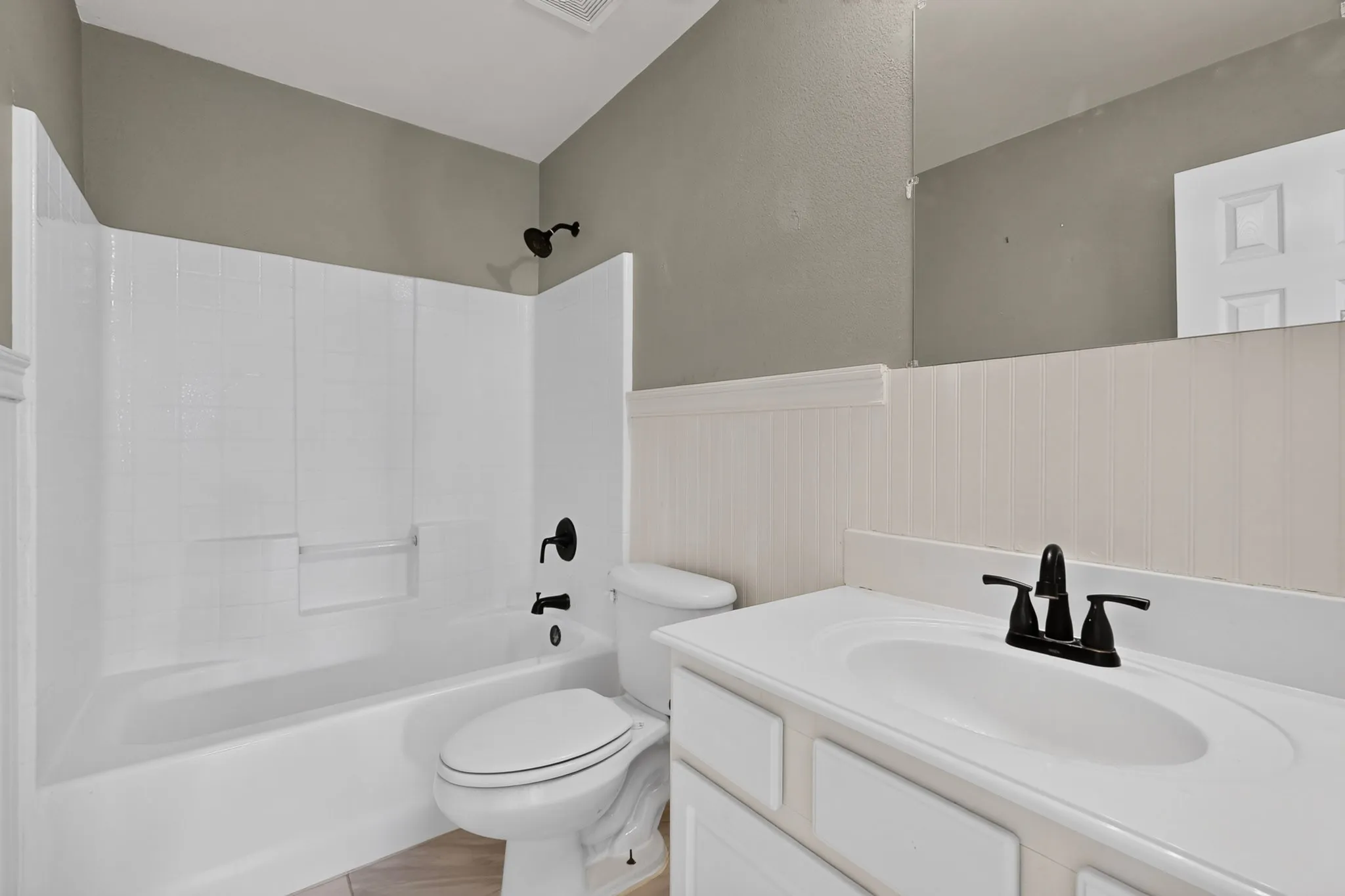 White bathtub with integrated shower, white vanity with integrated sink, matte black fixtures, and wainscoting detail