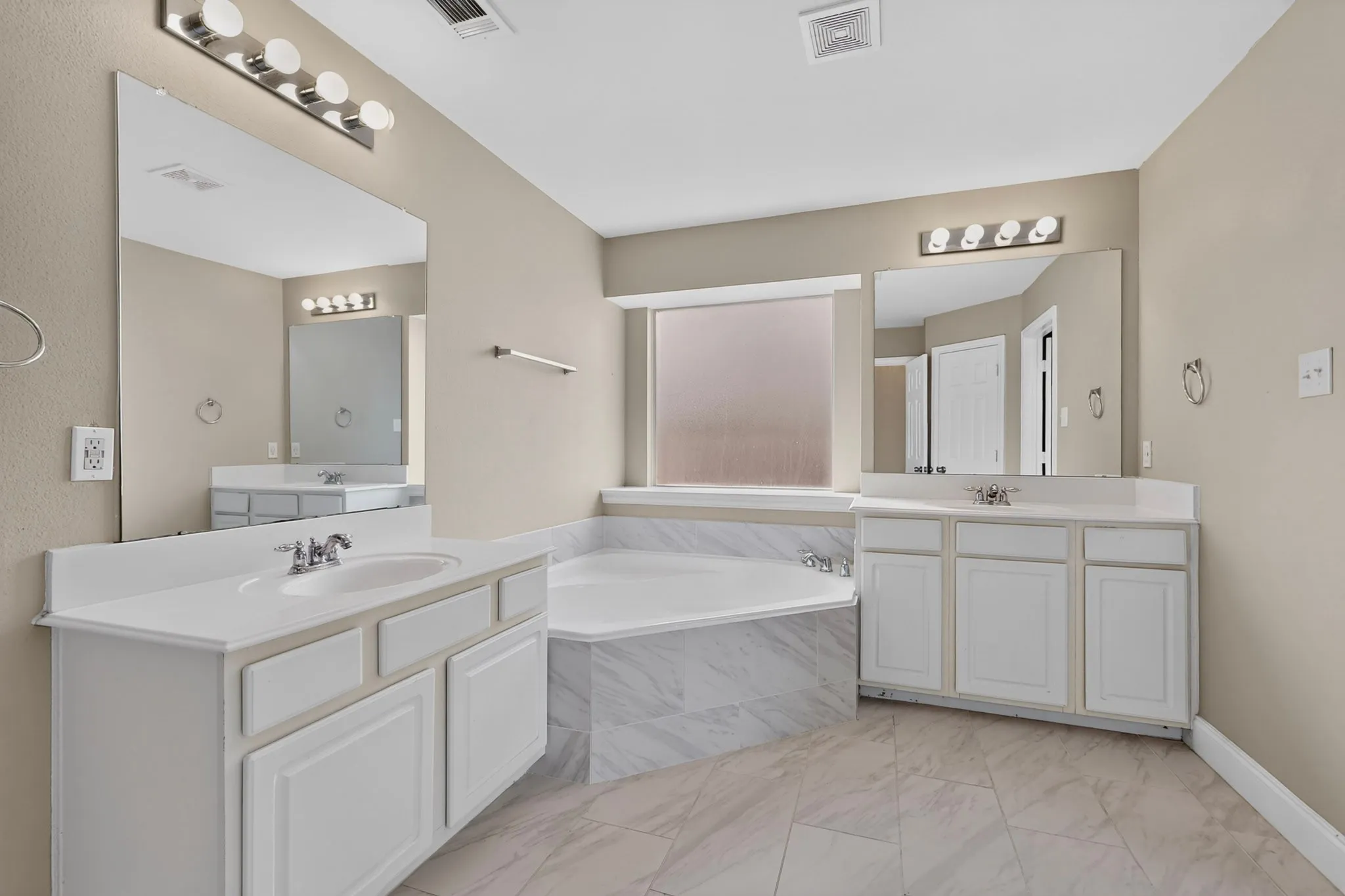 Dual vanity sinks with white cabinetry, built-in soaking tub with tile surround, large mirrors, and tile flooring