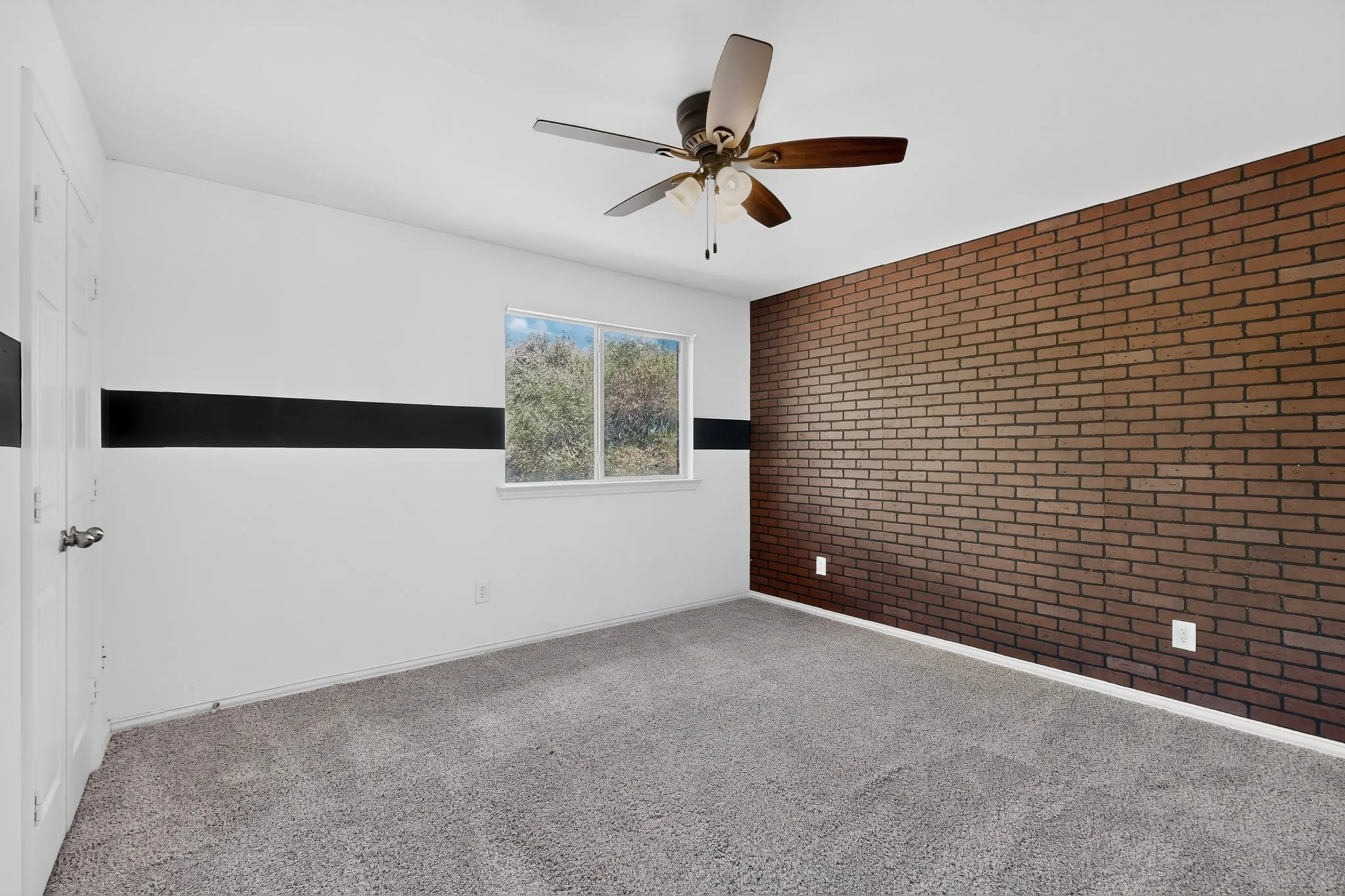 Accent brick wall, ceiling fan with integrated lighting, carpeted flooring, and a white-framed window