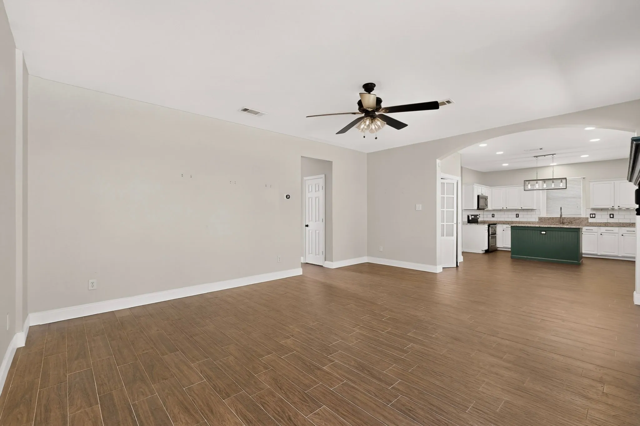 Wood-finish tile flooring throughout, ceiling fan with light fixture, arched doorway, and built-in white cabinetry with a dark green island in the kitchen