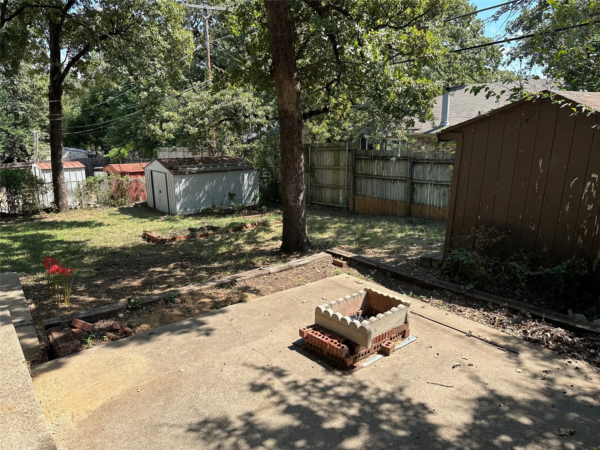 View from back patio of fenced backyard featuring 2 storage sheds and a paved area with custom firepit