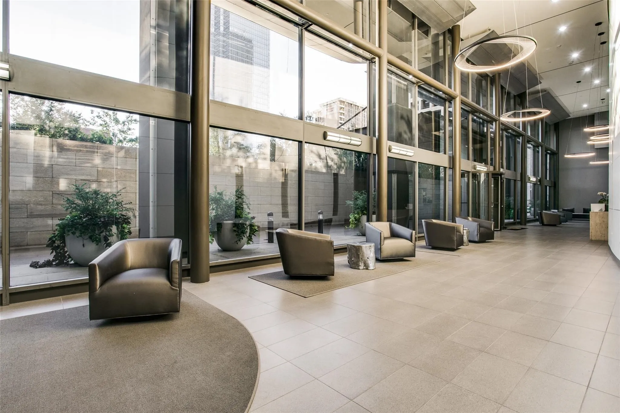 Expansive lobby featuring a tile floor, full-height windows, circular overhead lighting, and an exterior courtyard with planted greenery