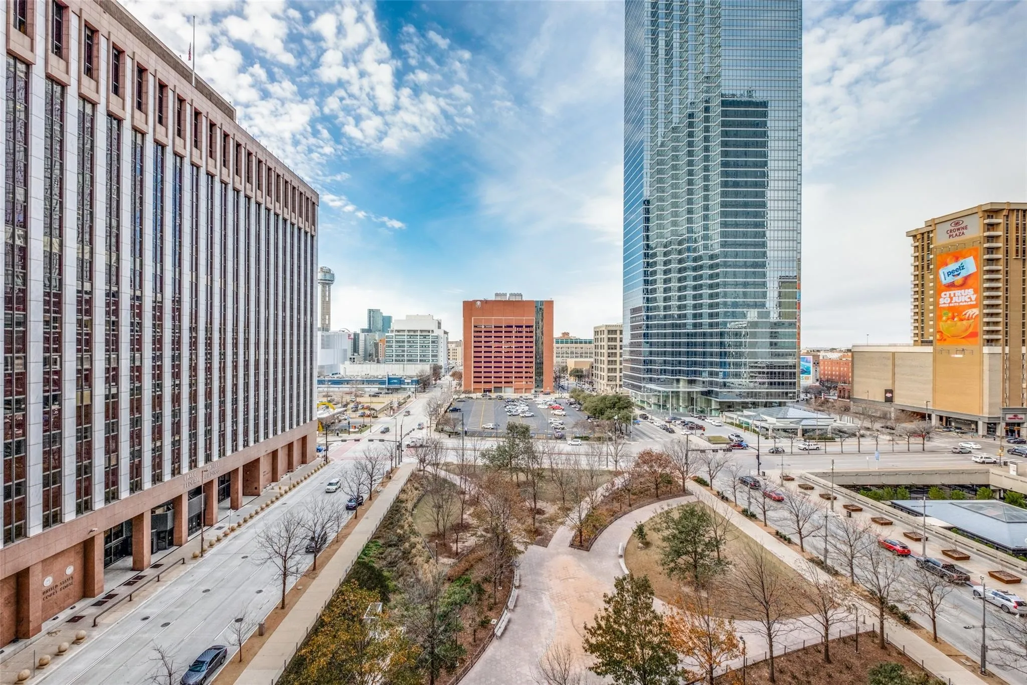 Urban landscape featuring a modern glass skyscraper, a multi-story commercial building, a structured city park with walkways, and surrounding street infrastructure