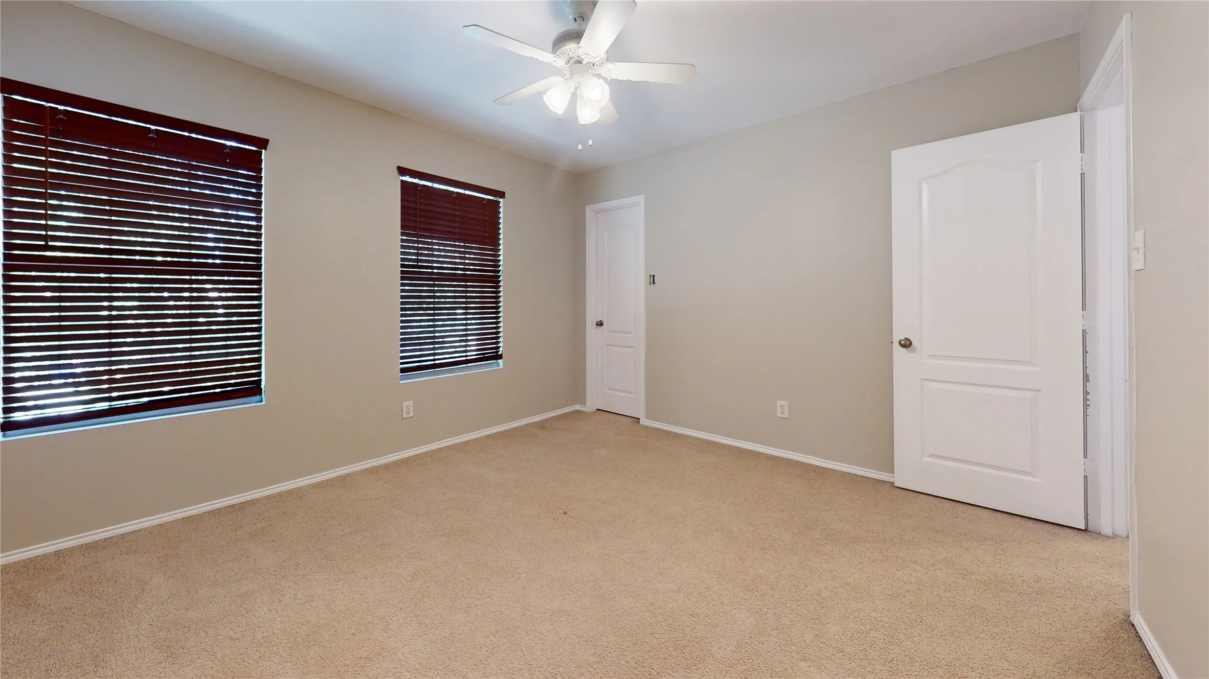 Spacious room with neutral wall tones, two windows with blinds, light-colored carpeting, and a ceiling fan