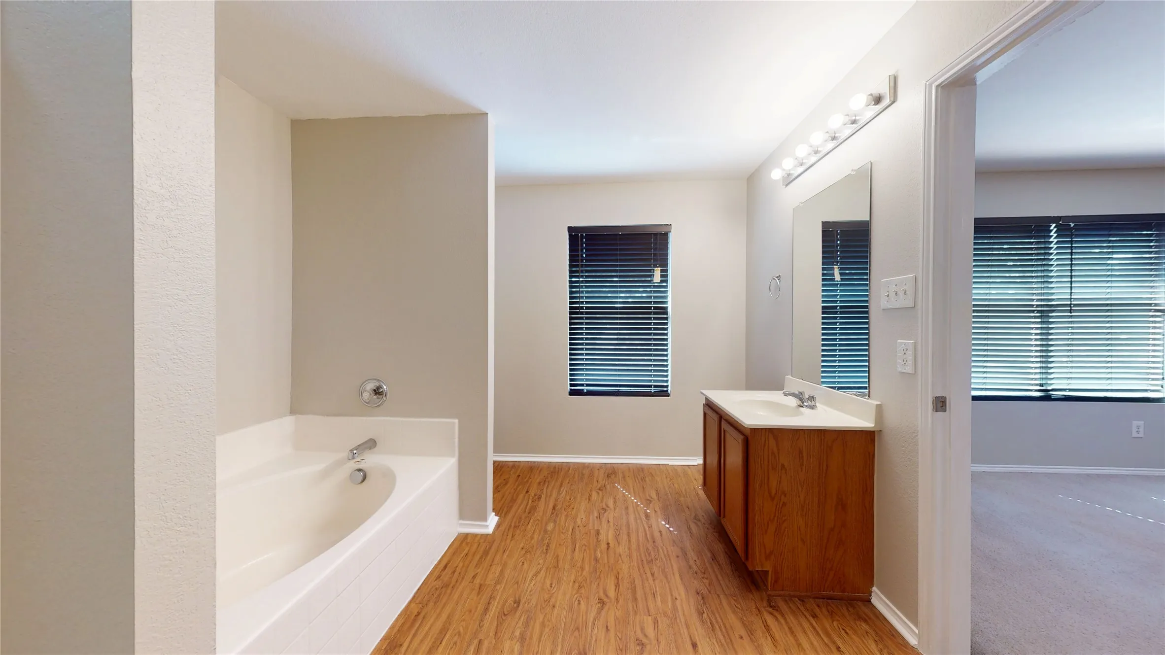 Soaking tub with chrome fixtures, wood-finish flooring, a wood vanity with a white countertop, and a vanity mirror with overhead lighting