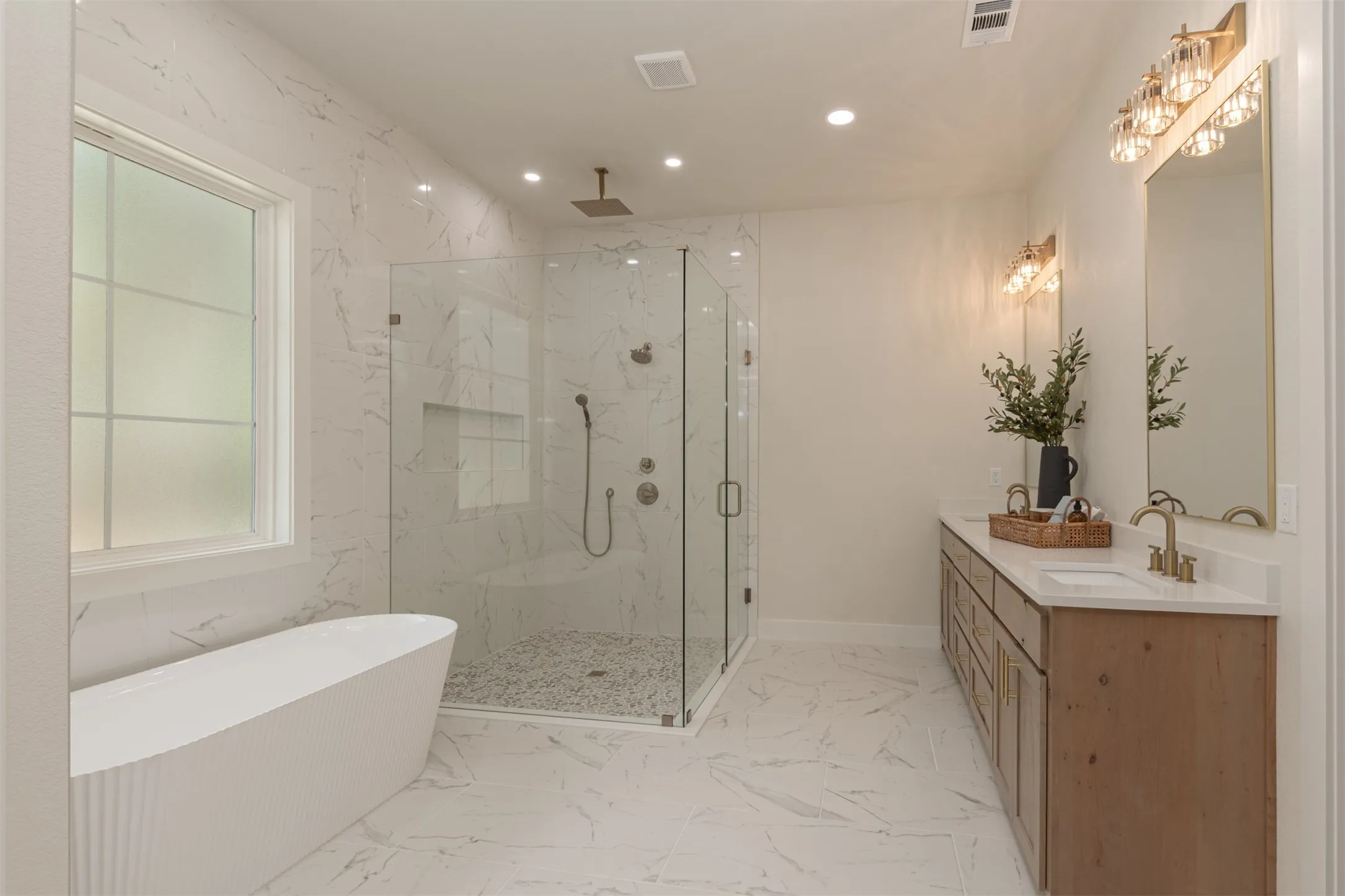 Freestanding soaking tub, frameless glass shower enclosure with rain shower head, dual vanity with wood-finish cabinetry, quartz countertops, and marble-look tile flooring