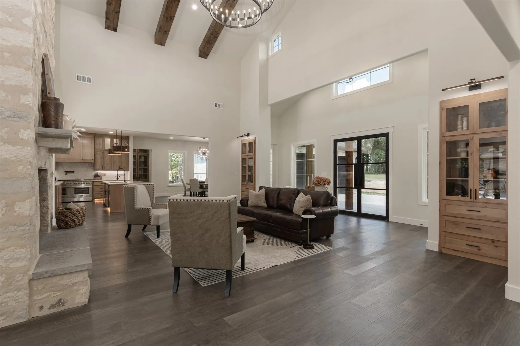 Vaulted ceiling with exposed wood beams, stone-clad fireplace with a hearth, wood-finish flooring, built-in display cabinetry, and black-framed double doors