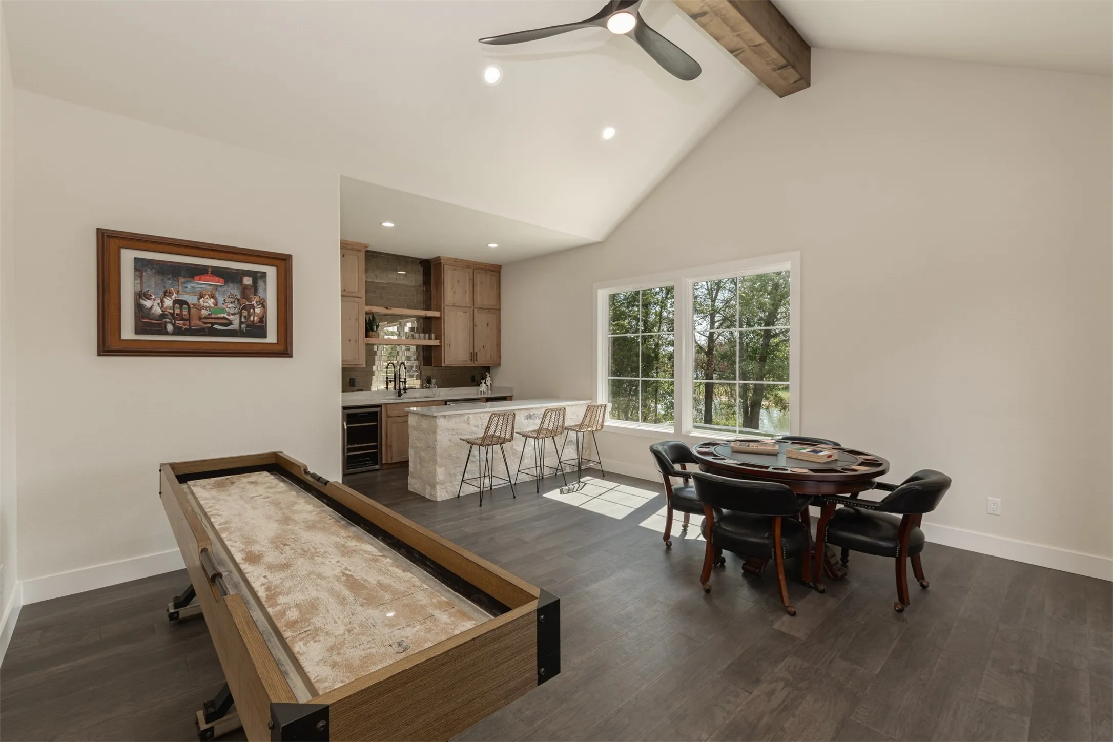 Vaulted ceiling with exposed wood beam, wood-finish flooring, built-in wet bar with stone-clad island, dual windows, and recessed lighting