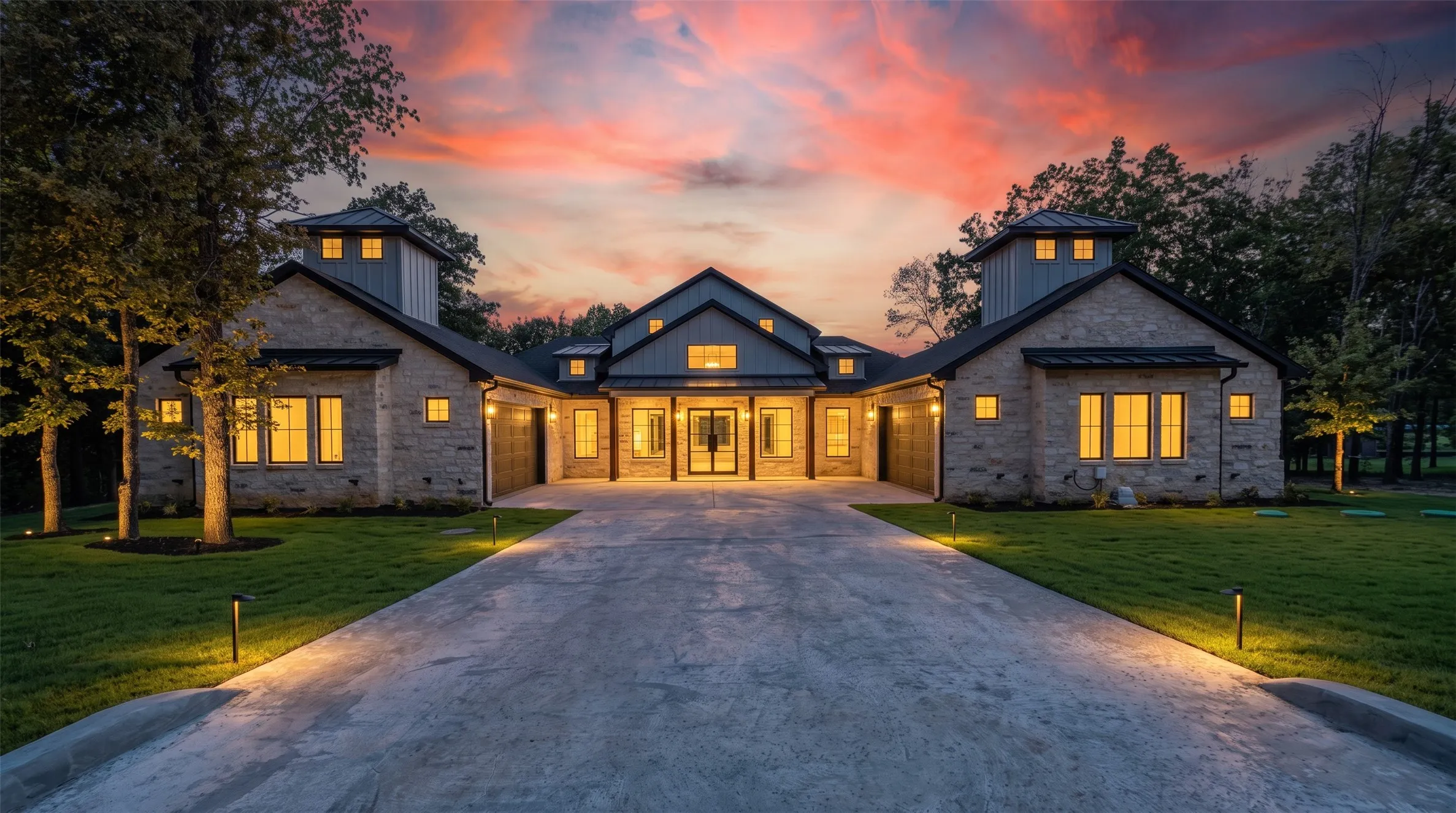 Stone facade with dark trim accents, multiple attached garages, a long concrete driveway, mature trees, and integrated landscape lighting