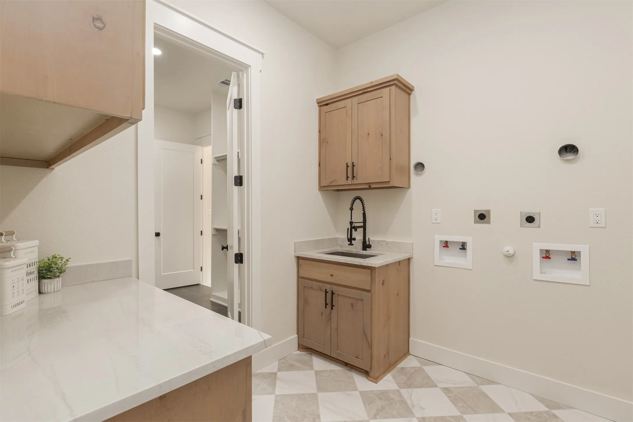 Utility room featuring checkered tile flooring, light wood-finish cabinetry, a sink with a matte black faucet, white countertops, and a built-in pantry