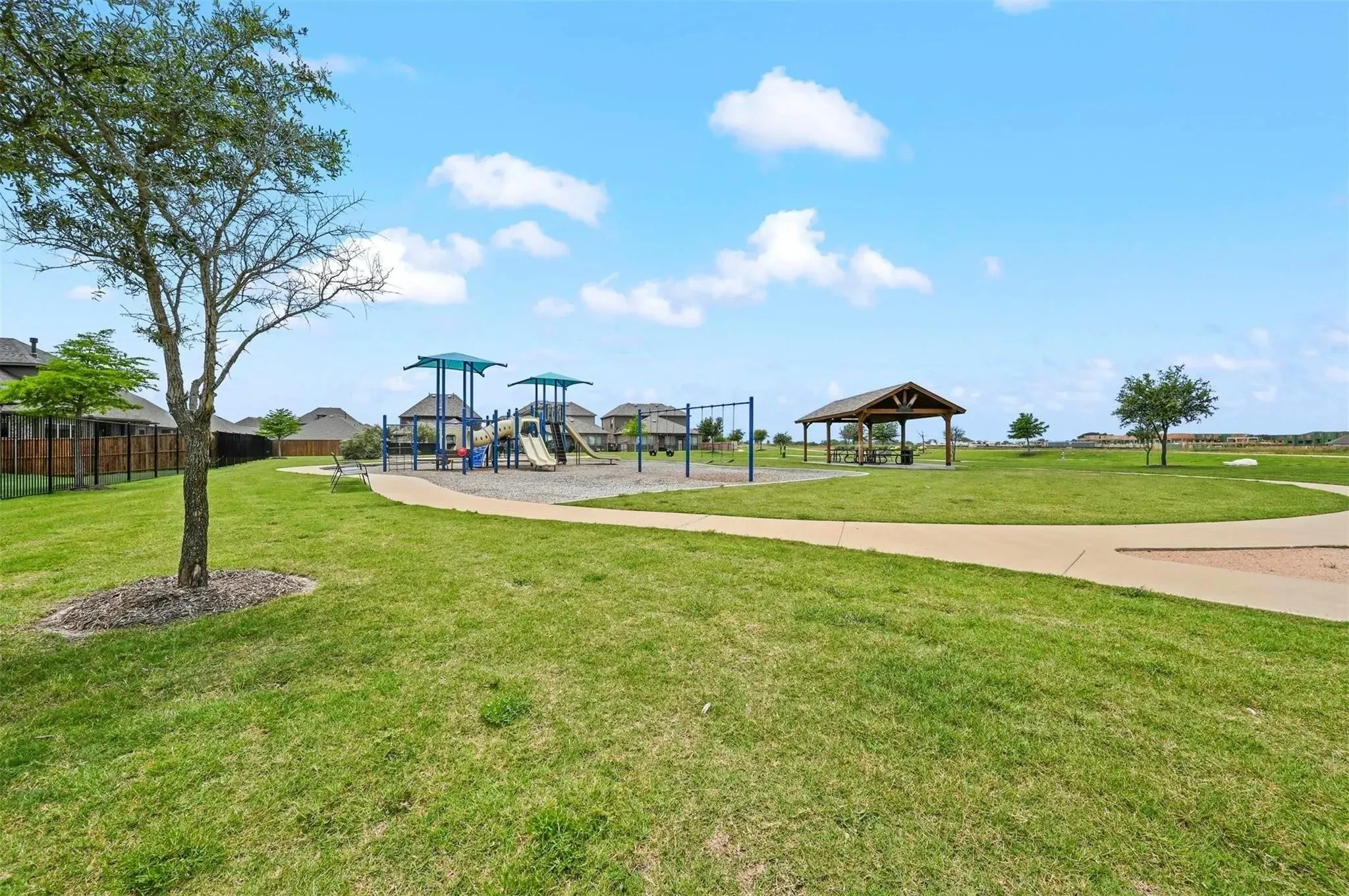 Expansive green space featuring a playground with shade structures, a covered picnic area, and paved walking paths