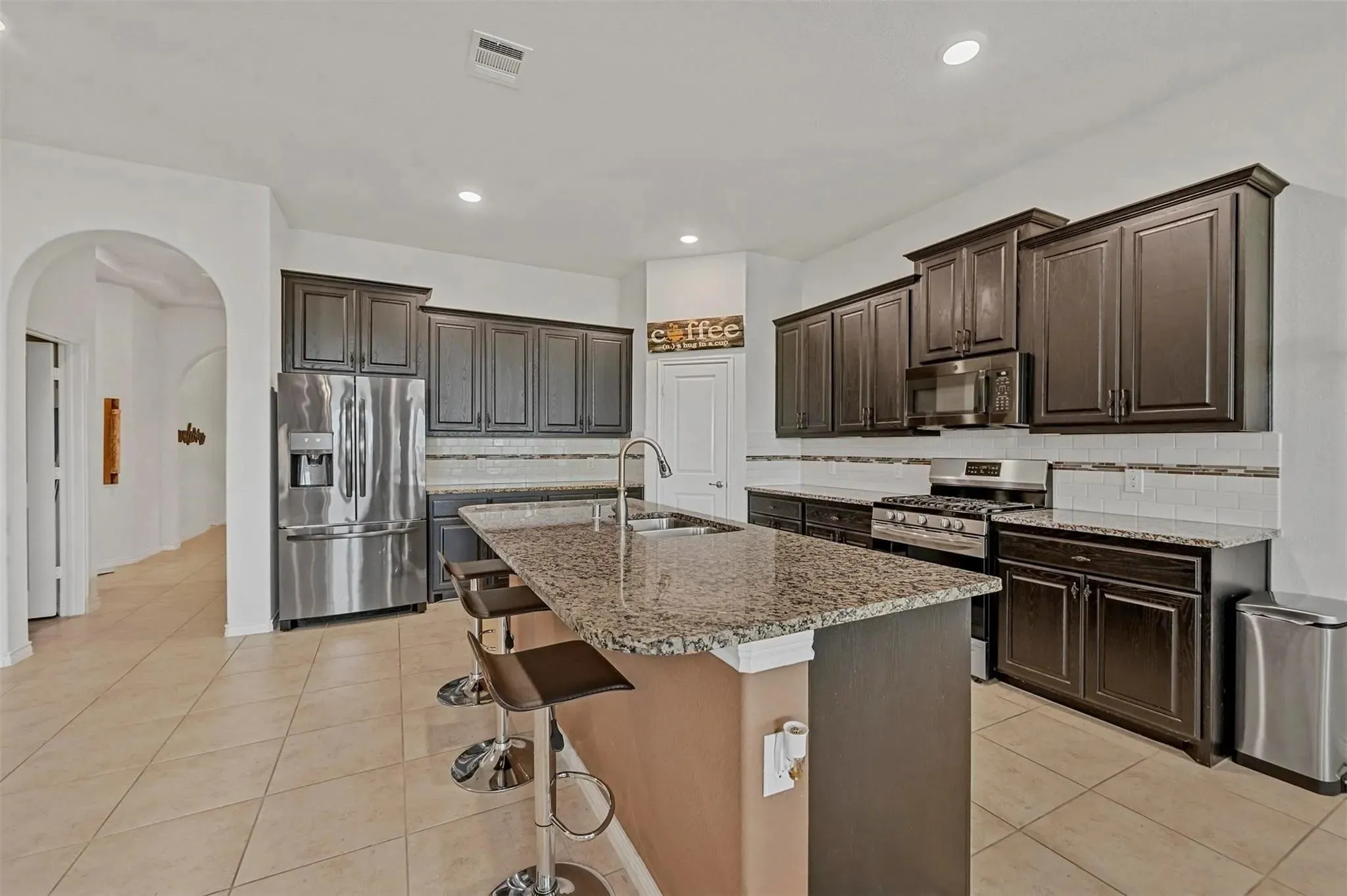 Kitchen featuring dark wood-finish cabinetry, stainless steel appliances, granite countertops, a large kitchen island with a sink, and tile flooring