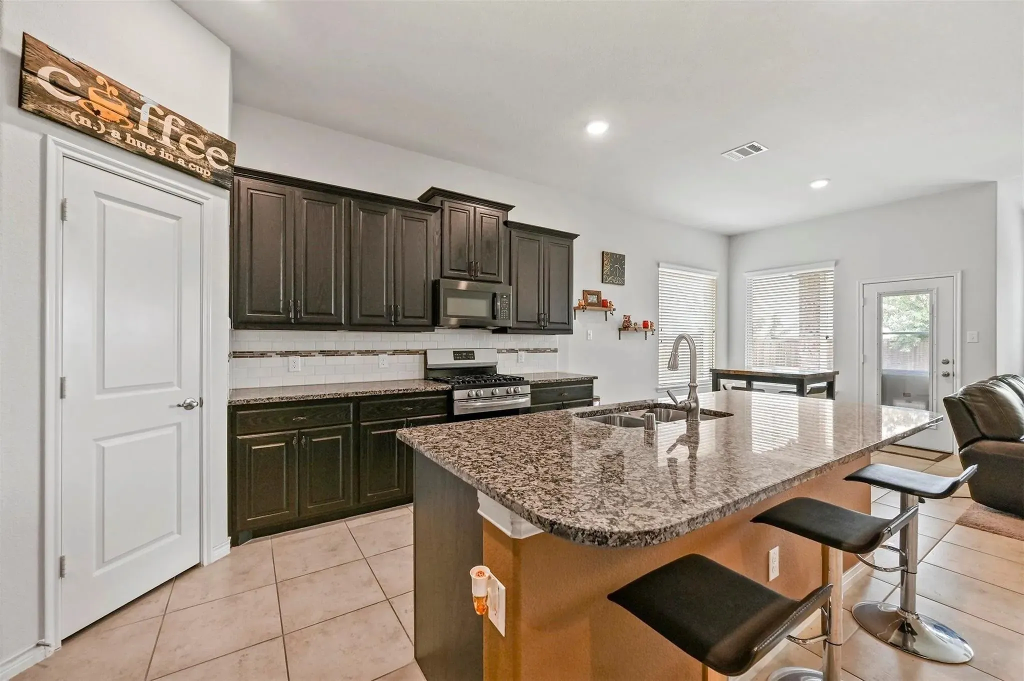 Kitchen featuring dark wood-finish cabinetry, stainless steel appliances, a multi-tone tile backsplash, and an island with a polished stone countertop and integrated sink