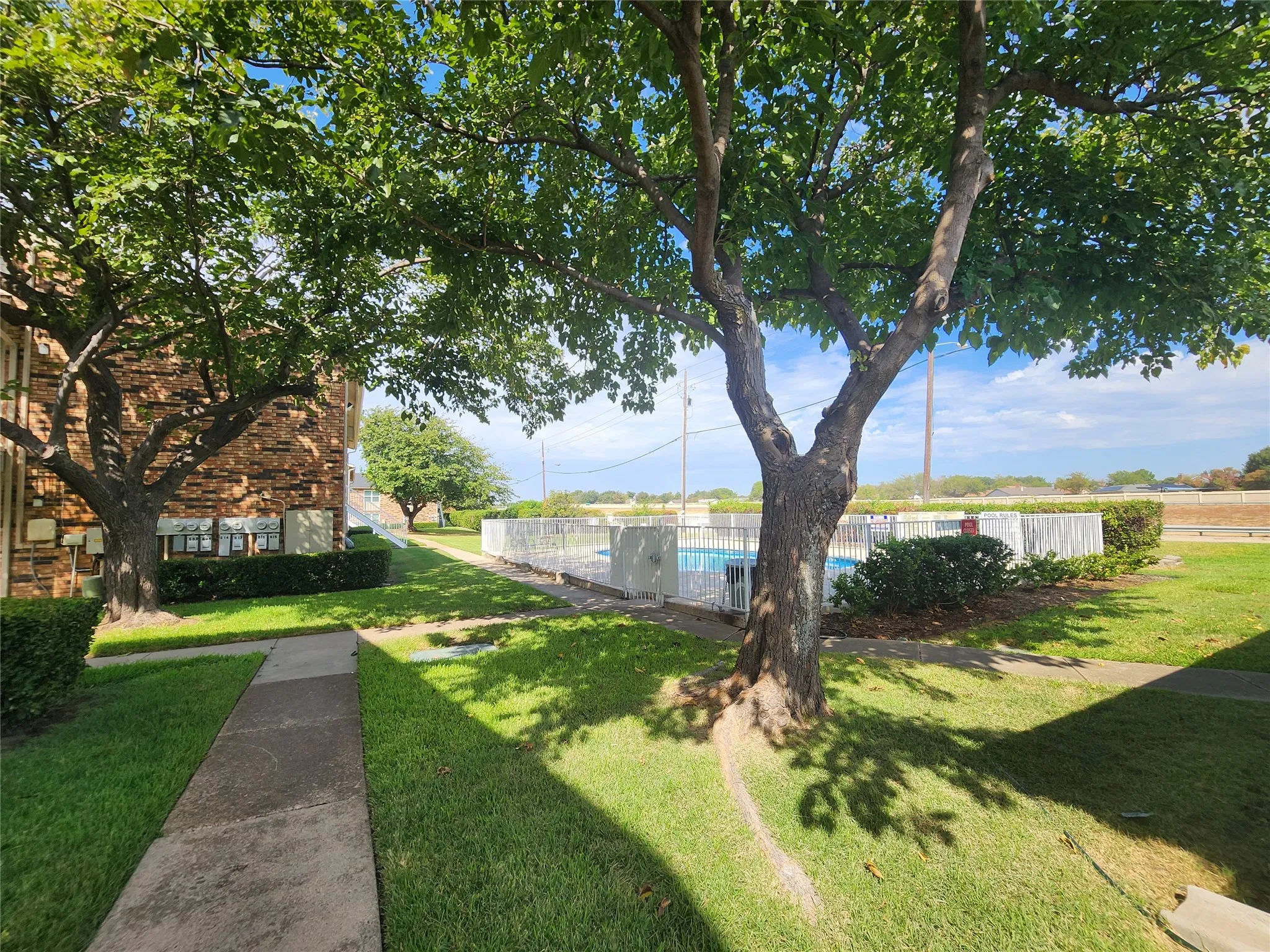 Lush green landscaping with mature trees, a paved walkway, exterior brick facade, and a fenced swimming pool