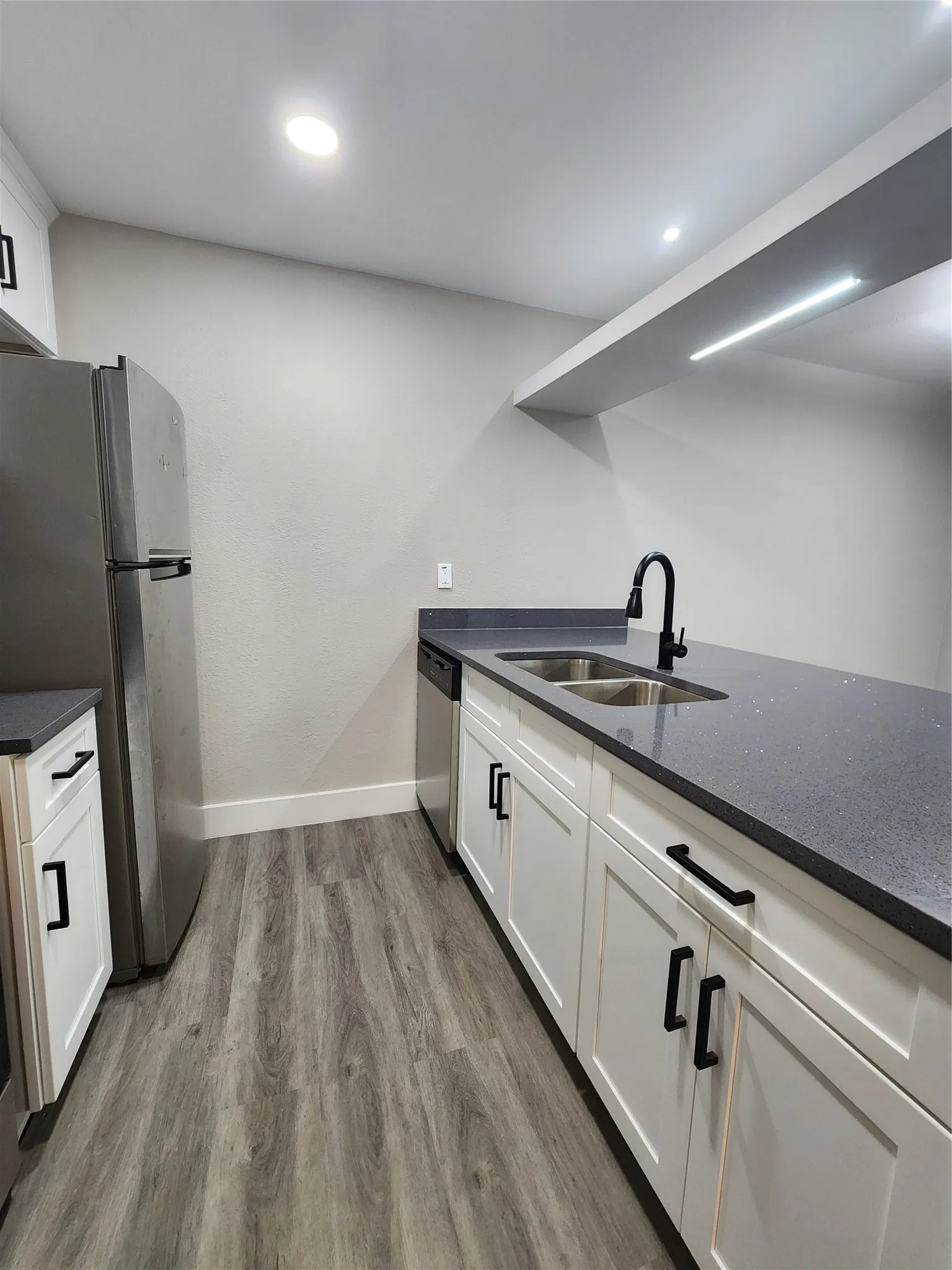 Kitchen featuring wood-finish flooring, white cabinetry with black hardware, dark countertops, a double basin stainless steel sink with a matte black faucet, and stainless steel appliances