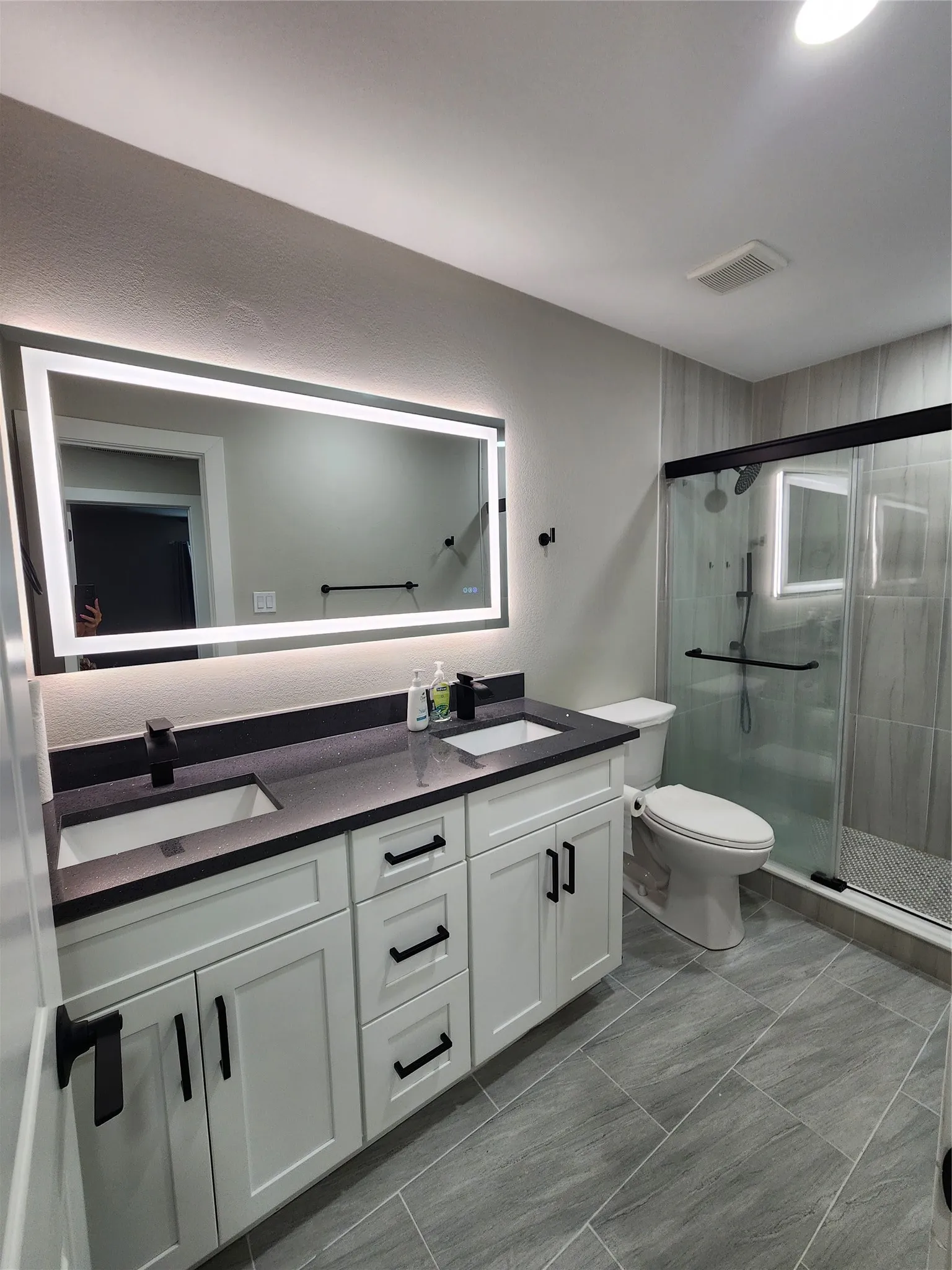 Dual sink vanity with dark countertops, white cabinetry, illuminated mirror, and a glass-enclosed shower featuring tiled walls