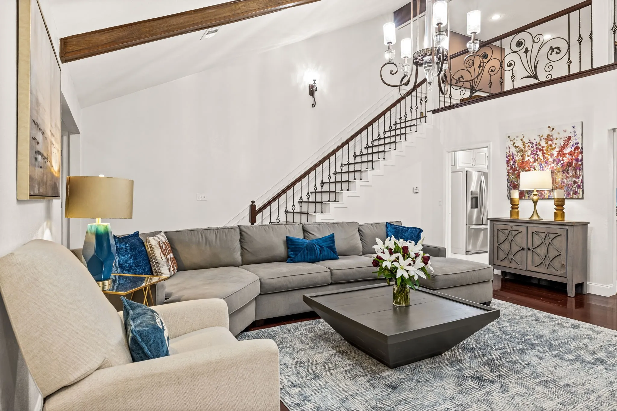 Vaulted ceiling with exposed wood beams, a prominent staircase with wrought iron balusters, a decorative upper-level railing, and dark wood-finish flooring