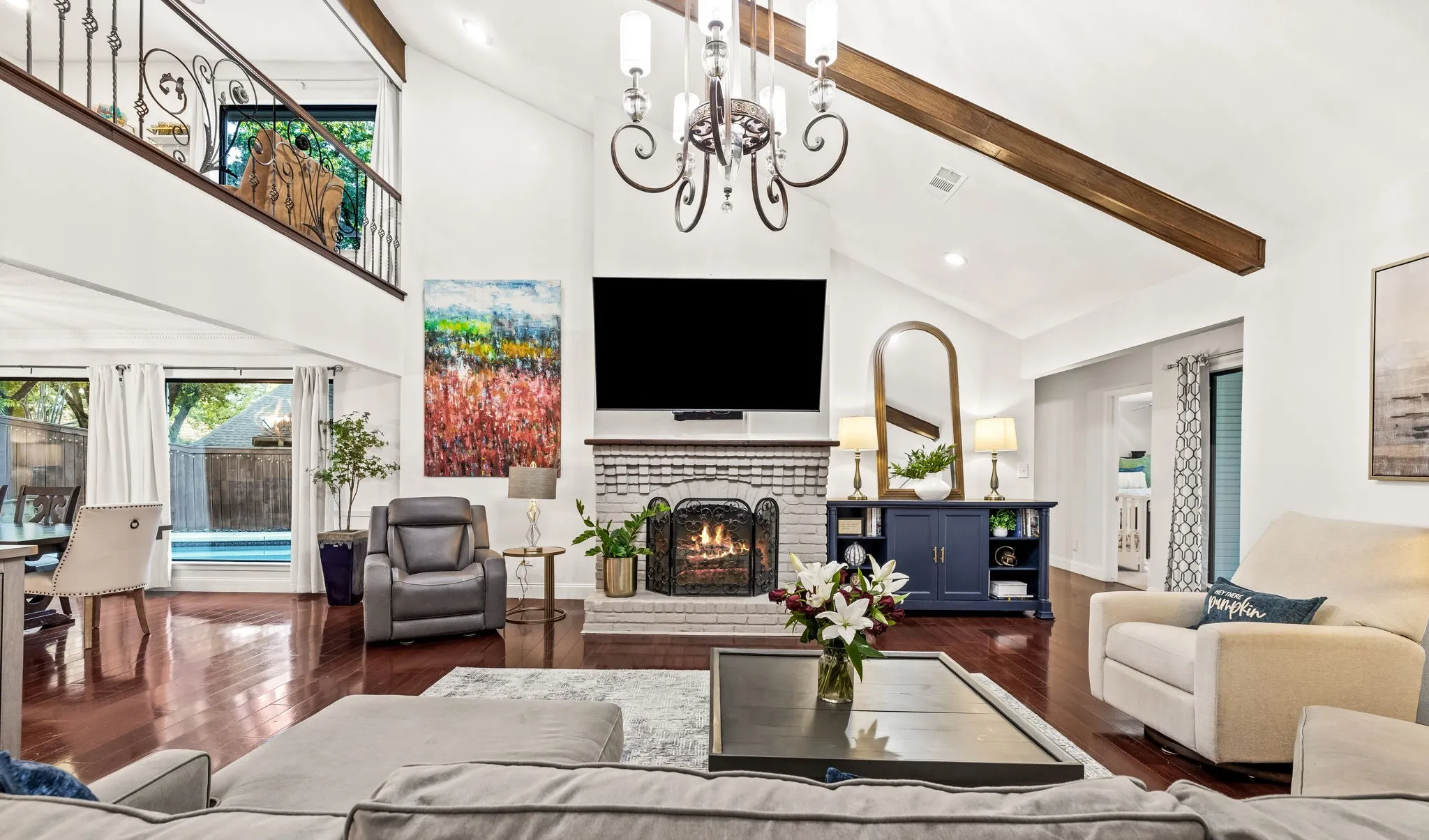 Vaulted ceilings with exposed wood beams, a brick fireplace, wood-finish flooring, and a second-story interior balcony with wrought iron railing