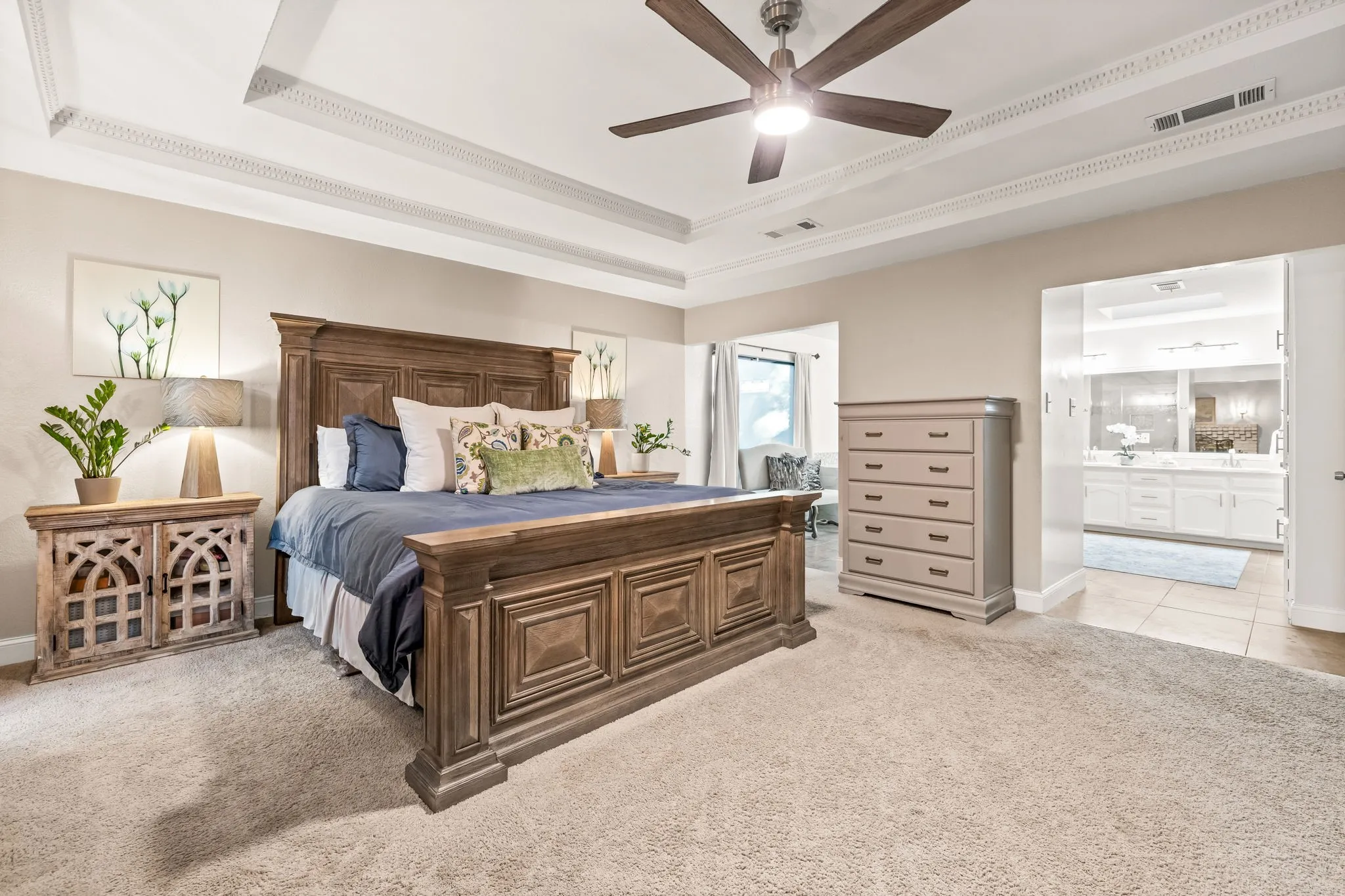 Tray ceiling with decorative molding, ceiling fan with integrated lighting, neutral wall tones, carpeted flooring, and an adjoining space with dual vanity