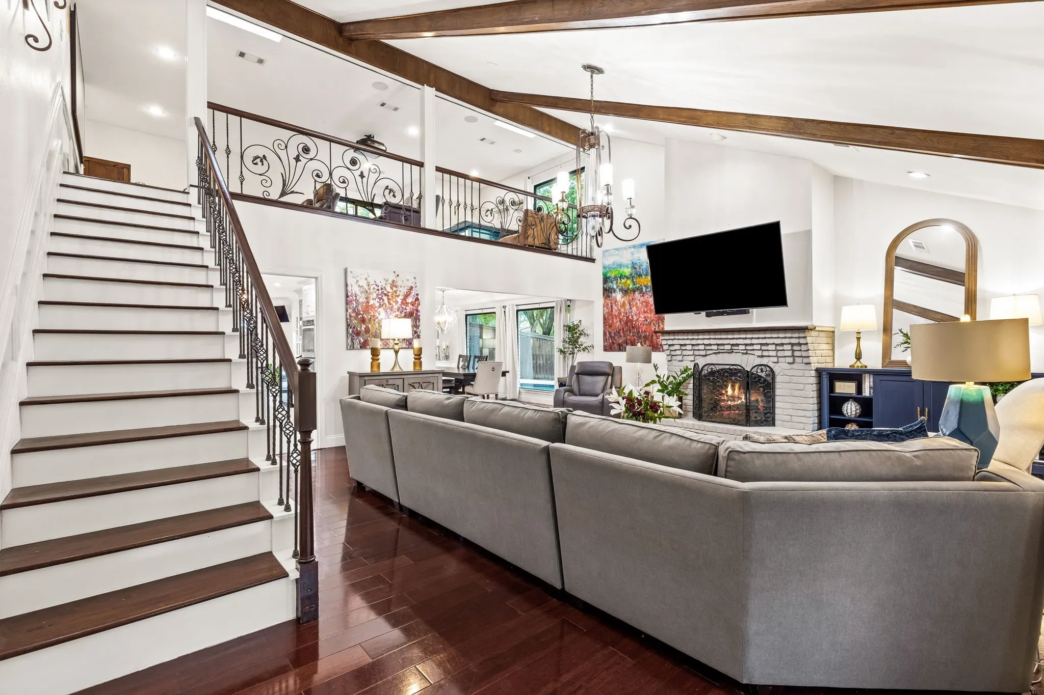 Vaulted ceiling with exposed wood beams, wood-finish flooring, brick fireplace, wrought iron staircase, and mezzanine balcony