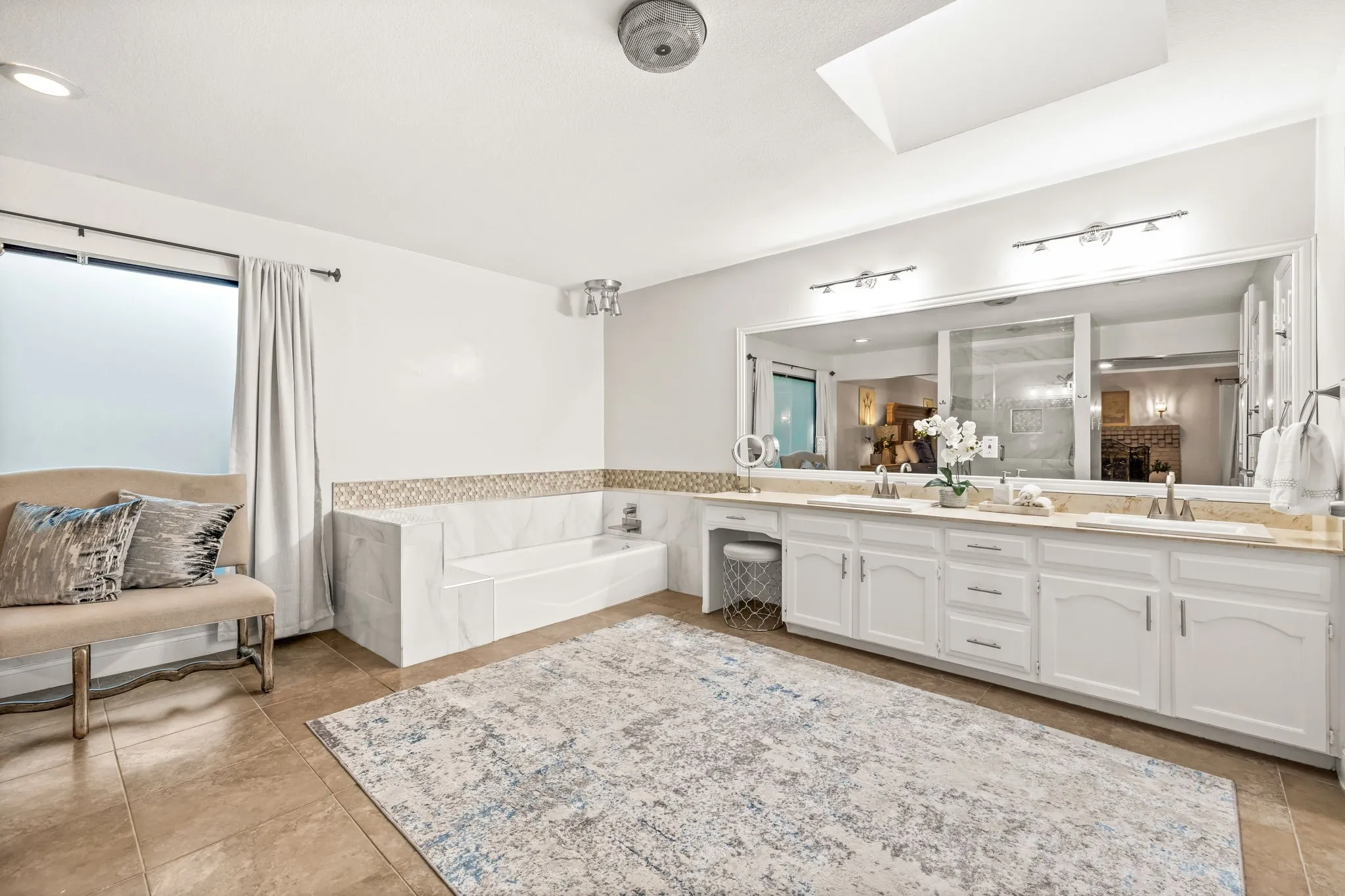 Spacious bathroom with dual vanity, integrated soaking tub, large wall mirror, skylight, and tiled flooring