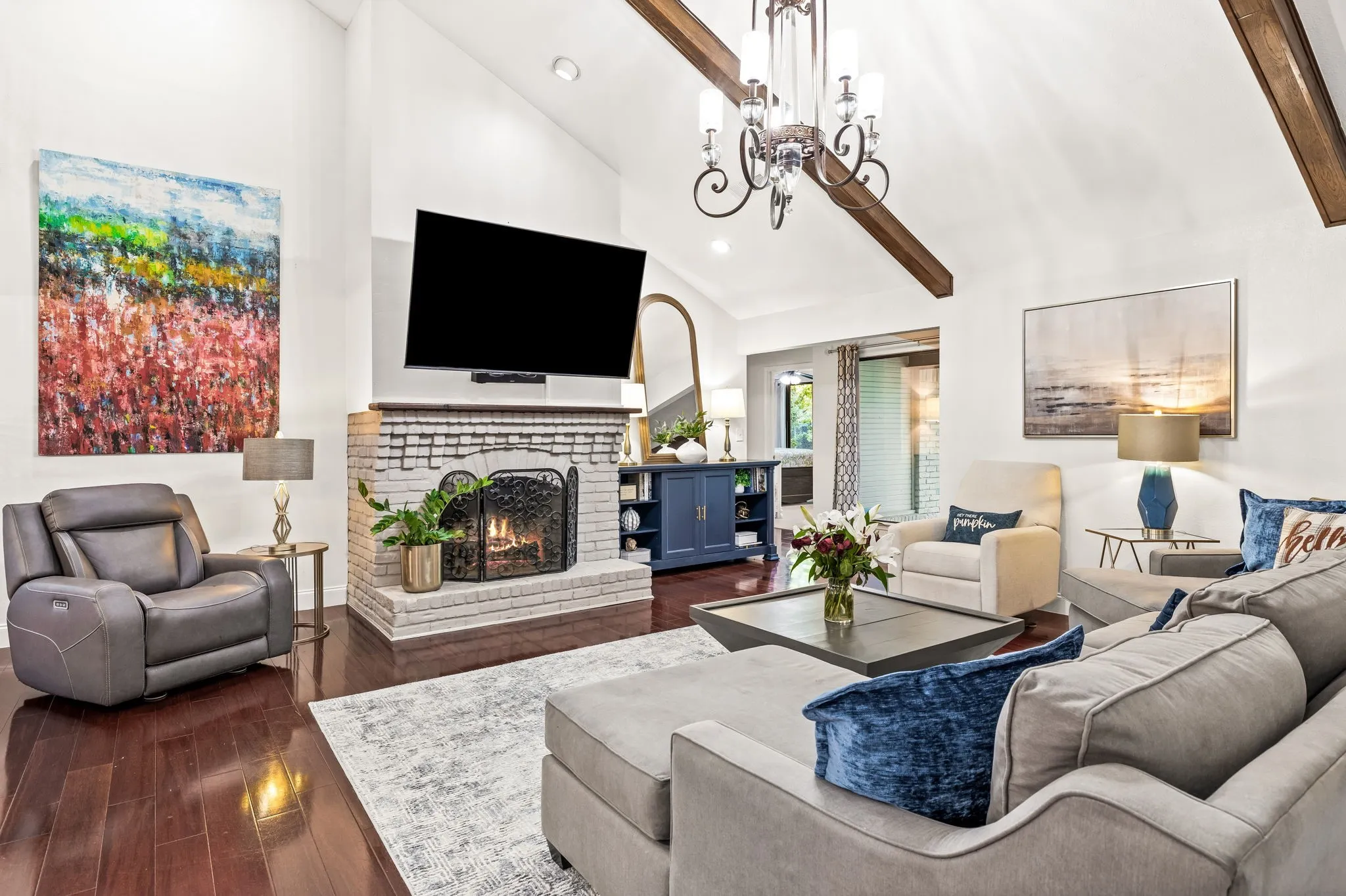 Vaulted ceiling with exposed wood beams, brick fireplace with metal screen, wood-finish flooring, and chandelier