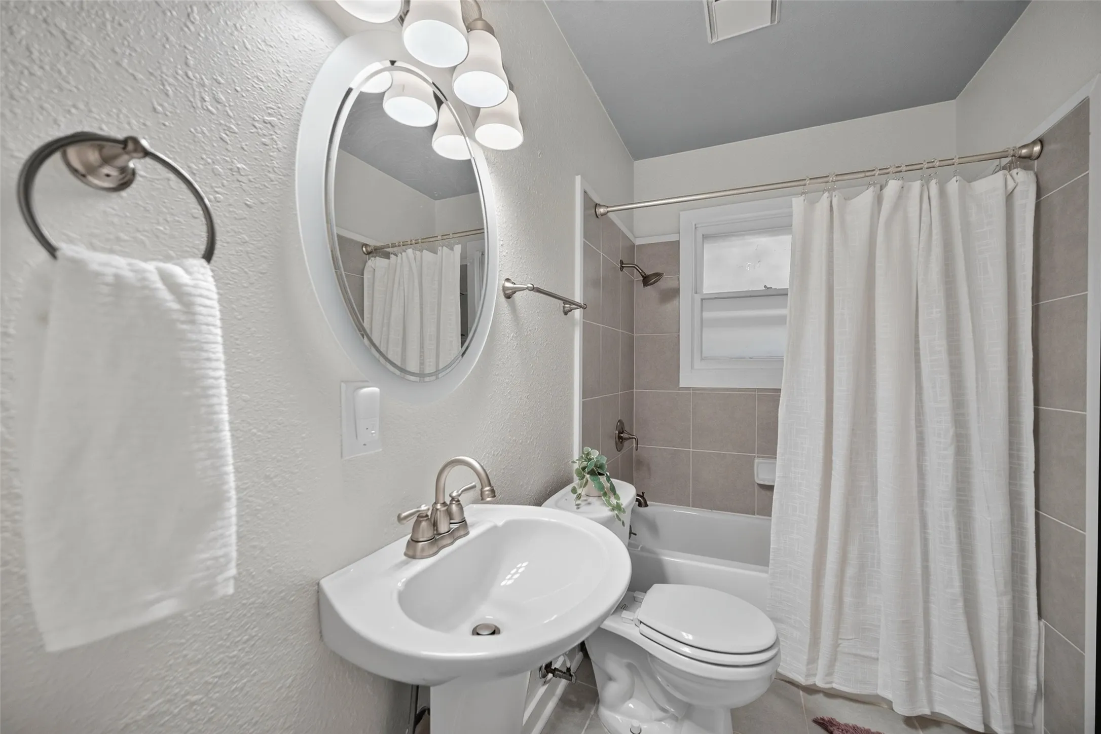 Pedestal sink with a brushed nickel faucet, oval framed mirror, sconce lighting, tiled shower surround, and a double-hung window