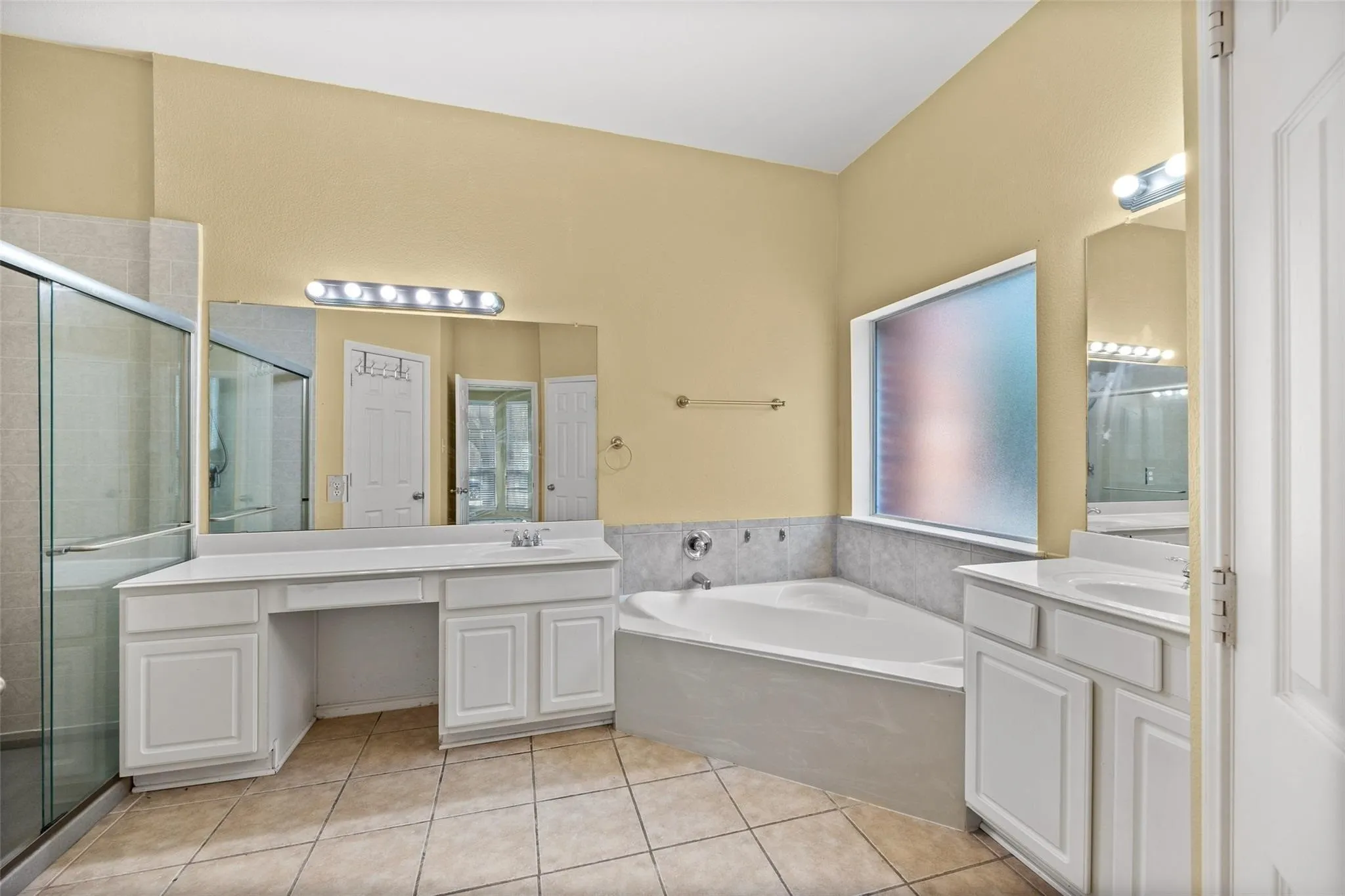 Dual vanity with integrated sinks, a built-in makeup counter, a large soaking tub, a tiled shower with a glass enclosure, and a privacy window