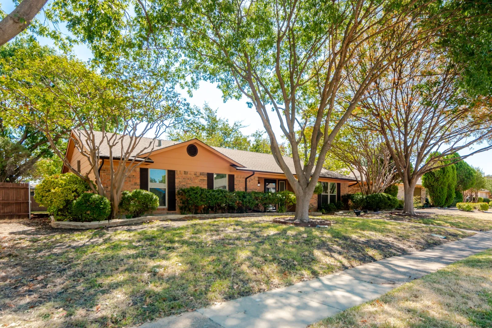 Brick exterior with dark shutters, mature trees, landscaped front yard, and a concrete sidewalk