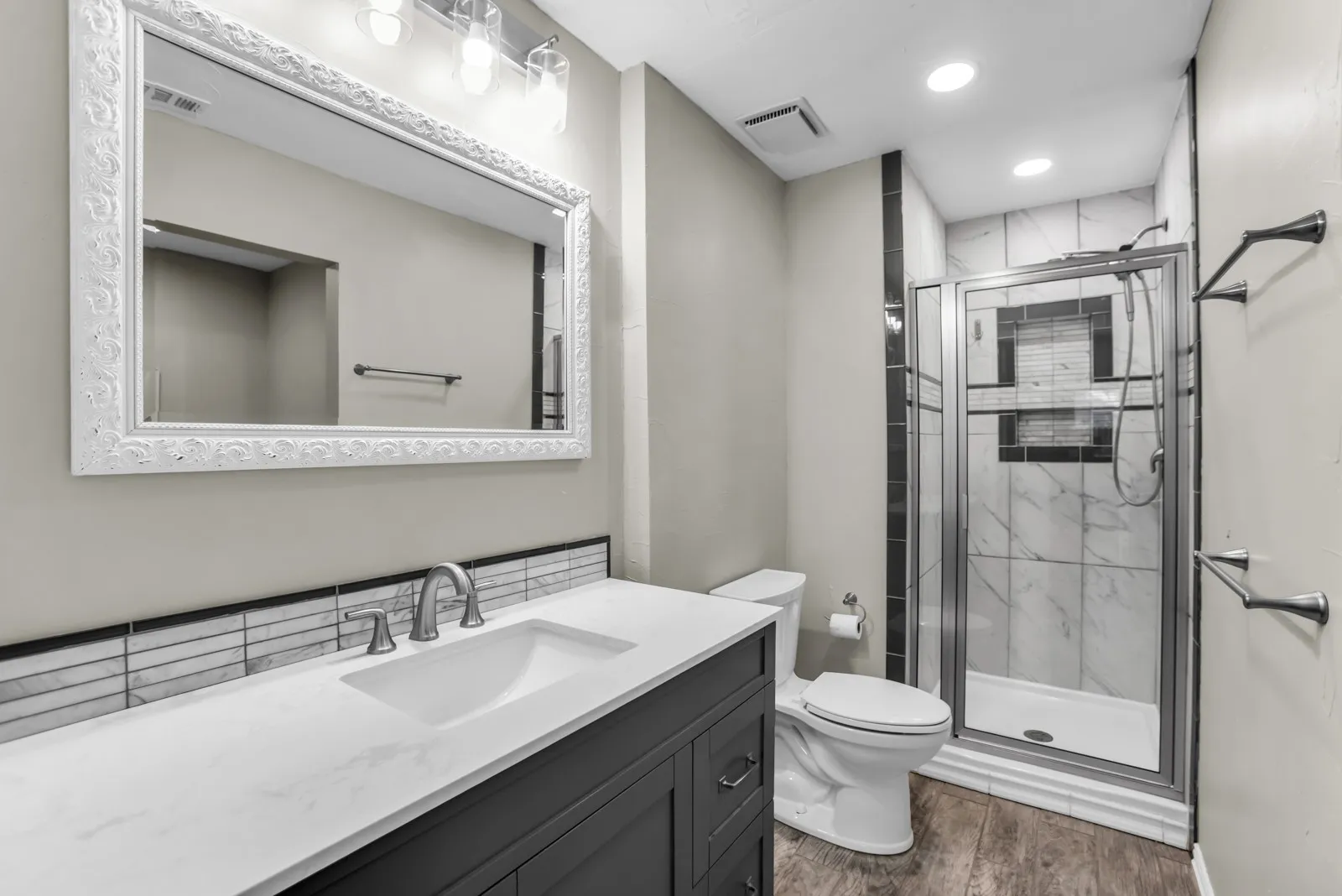 Vanity with a white countertop and integrated sink, framed mirror with overhead lighting, glass-enclosed shower with tiled walls, and wood-finish flooring