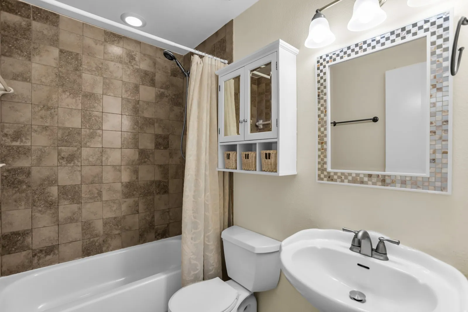 Bathtub with tiled surround, wall-mounted storage cabinet, mirror with mosaic tile frame, pedestal sink, and vanity light fixture