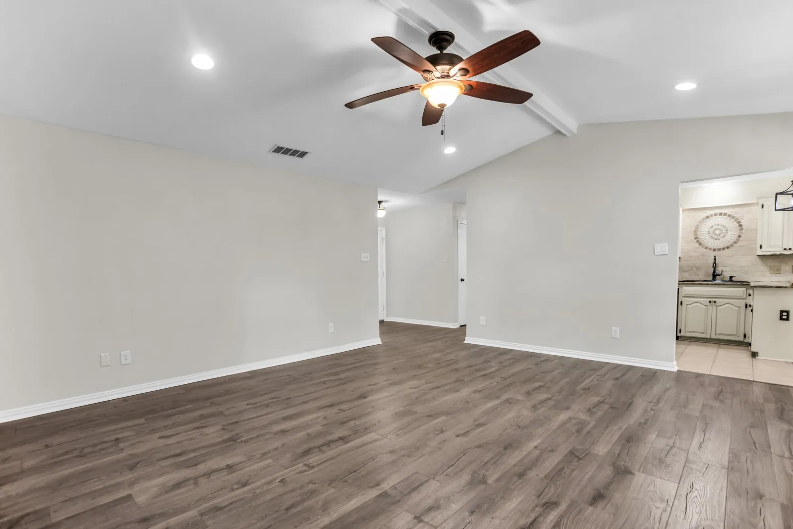 Vaulted ceiling with exposed beams, recessed lighting, wood-finish flooring, ceiling fan with integrated lighting, and a partial view of a kitchen with light-colored cabinetry