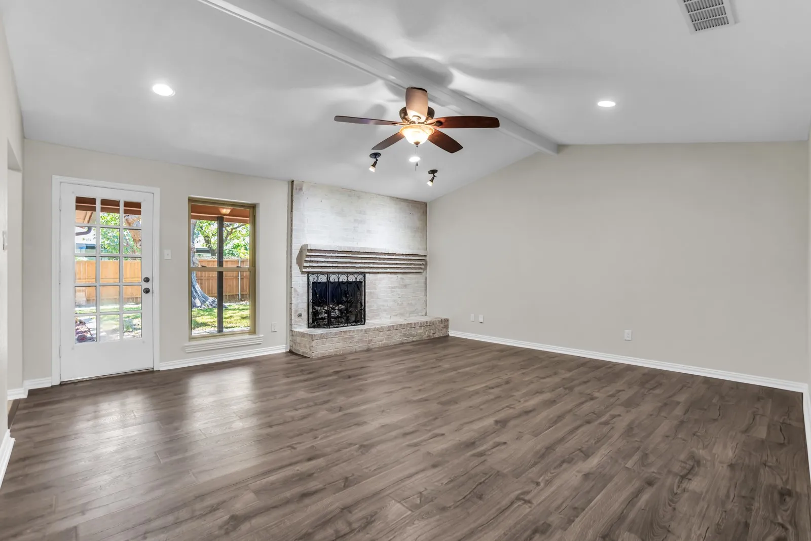 Vaulted ceiling with exposed beam, wood-finish flooring, white-painted brick fireplace, French door to backyard, and recessed lighting