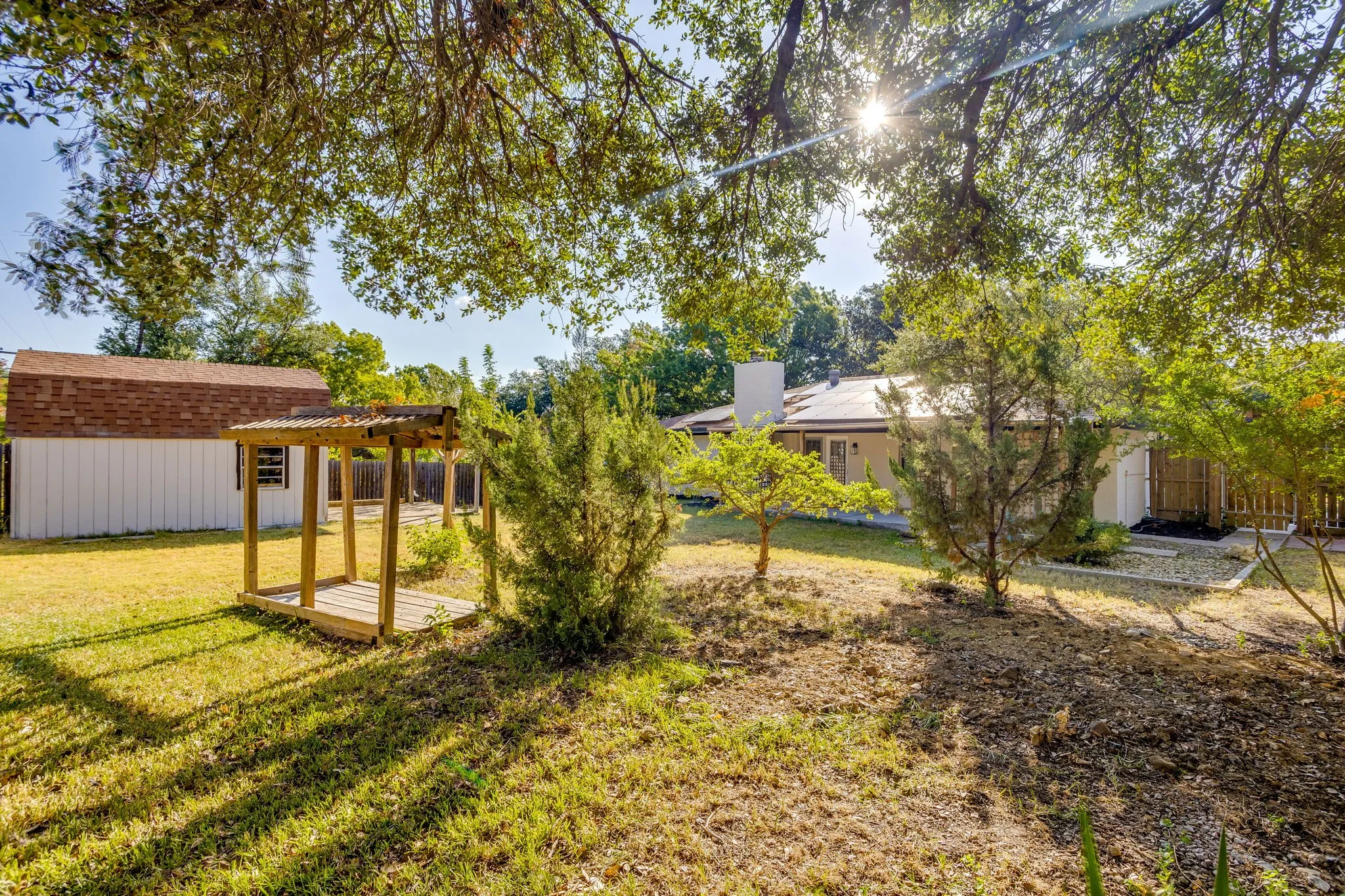 Expansive backyard featuring a detached storage shed with a shingle roof, a wooden pergola, mature trees, and a gravel landscape bed