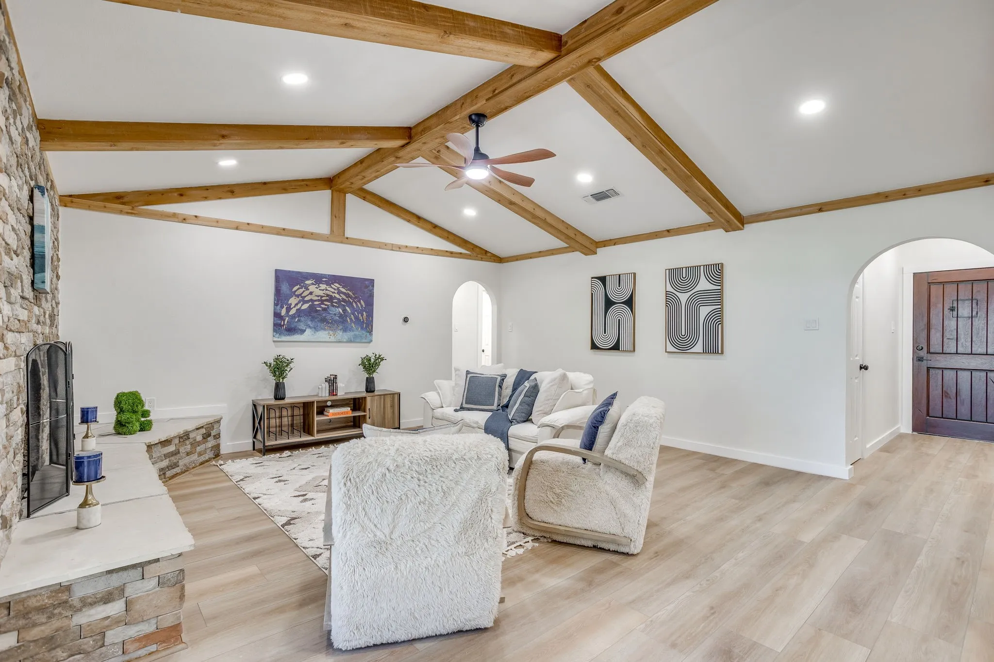 Vaulted ceiling with exposed wood beams, stone-clad fireplace with hearth, wood-finish flooring, recessed lighting, and arched doorway