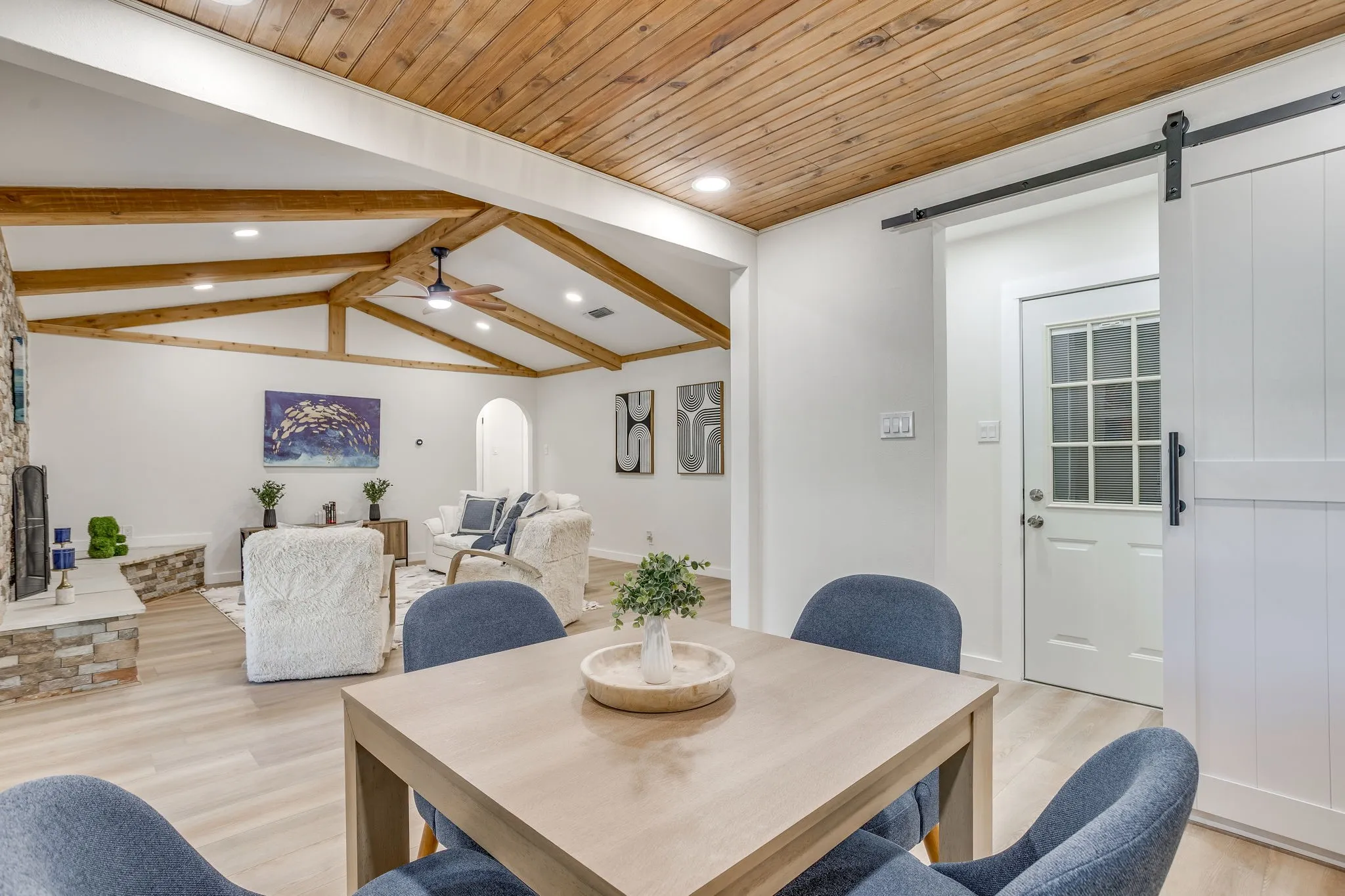 Vaulted wood-paneled ceiling with exposed beams, wood-finish flooring, white barn door, white paneled door with glass insert, and stacked stone fireplace surround