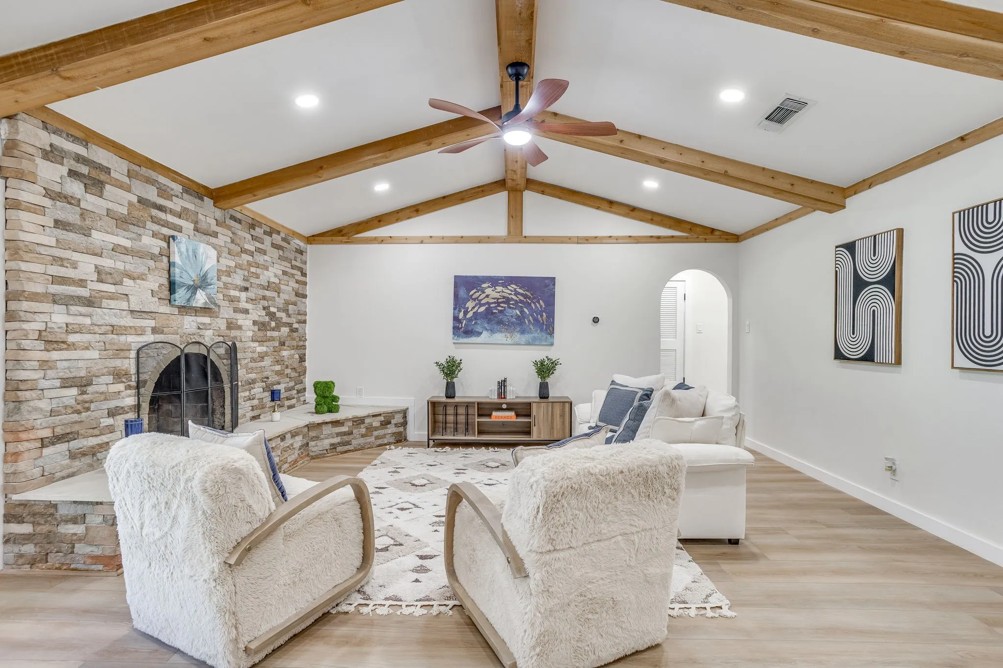 Vaulted ceiling with exposed wood beams, a stone-clad fireplace, wood-finish flooring, recessed lighting, and a ceiling fan