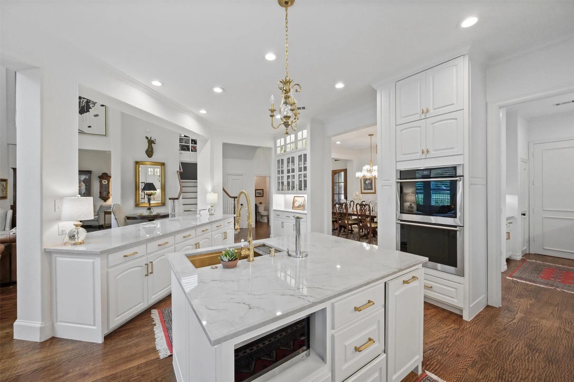 Gourmet kitchen featuring a substantial island with countertop seating, a gold-toned sink, double wall ovens, extensive white cabinetry, and wood-finish flooring