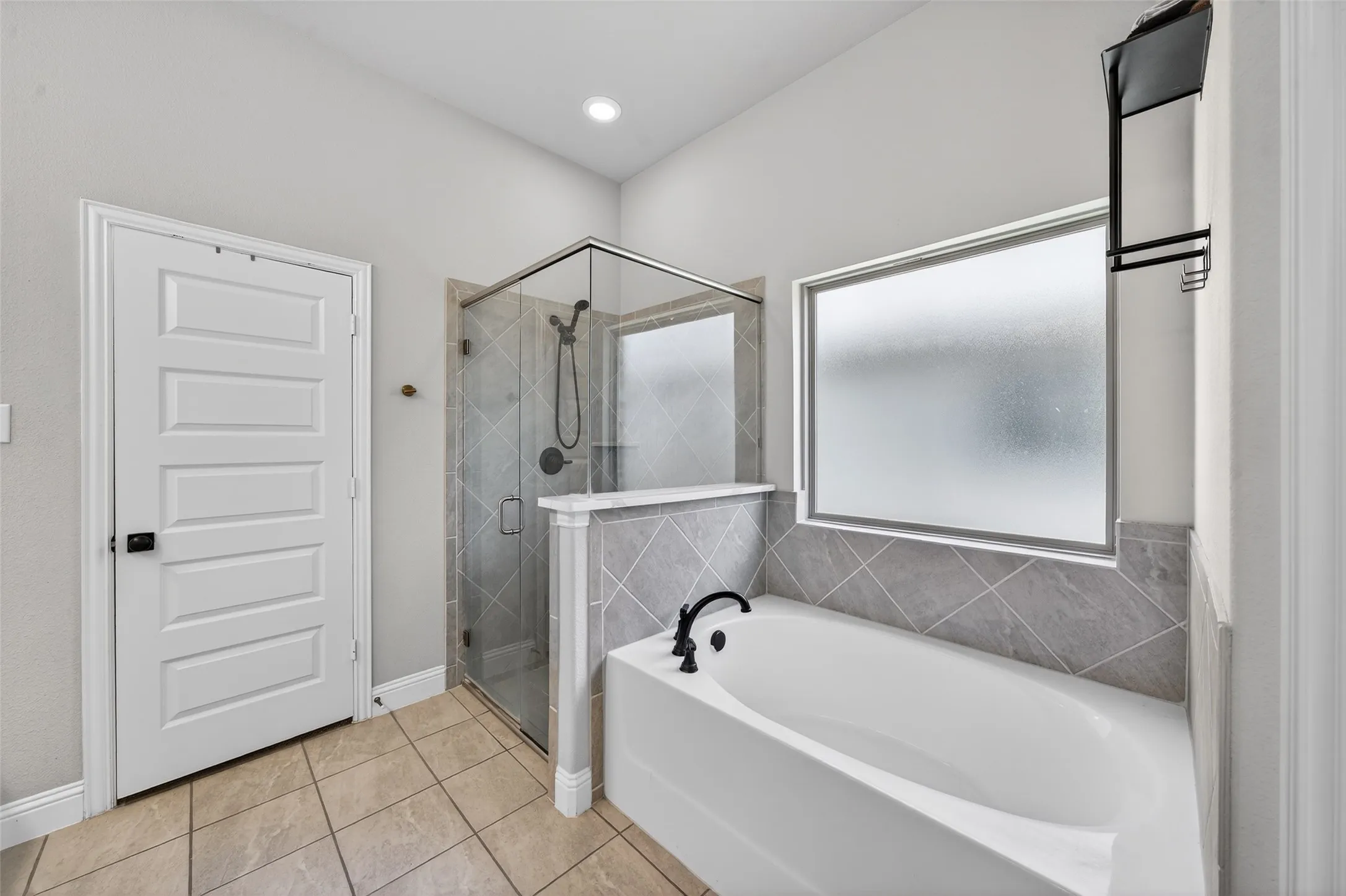 Tiled flooring, built-in soaking tub with dark bronze fixtures, glass-enclosed shower with subway tile surround, and a privacy window