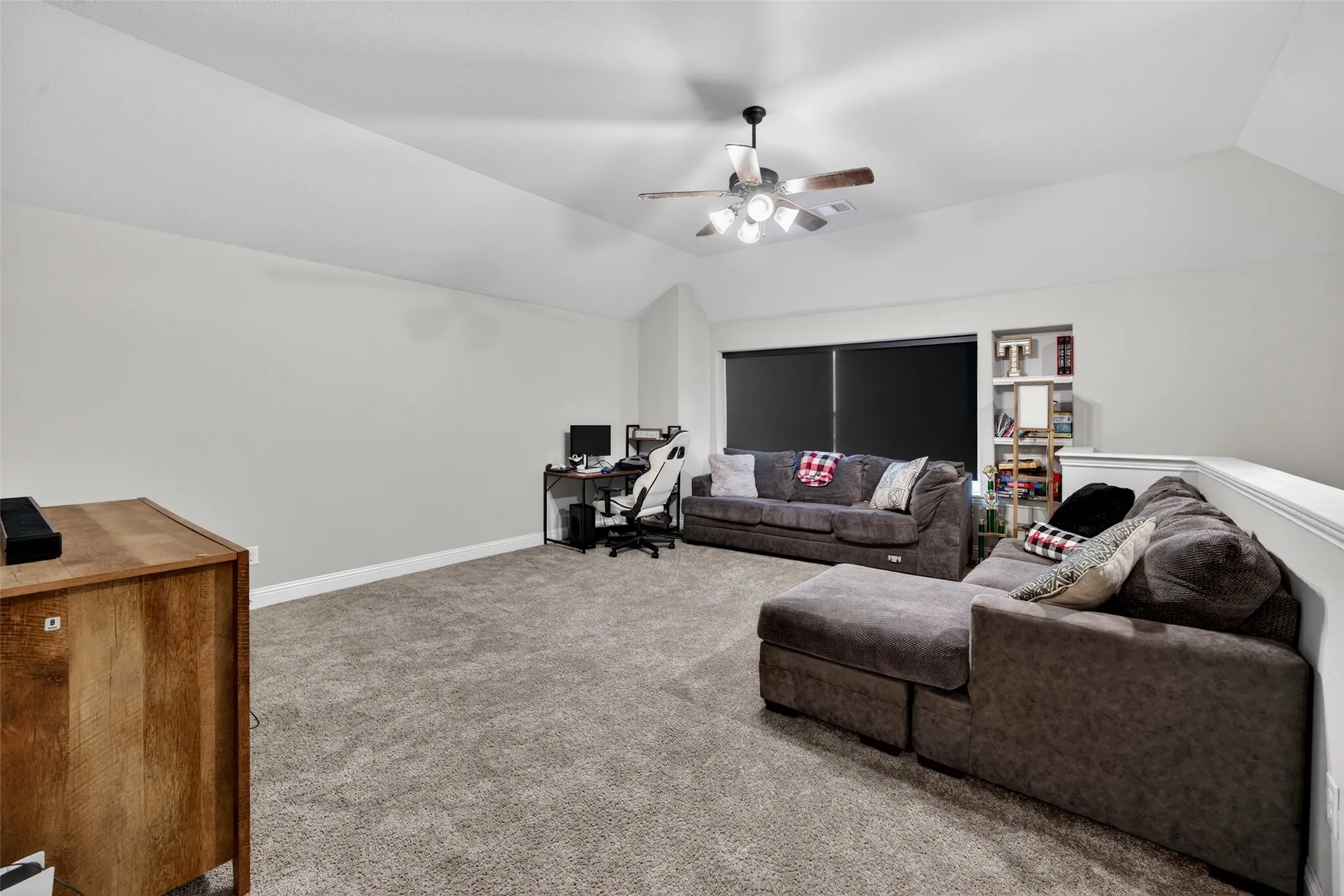 Carpeted room featuring a ceiling fan with integrated lighting, a window with roller shades, and built-in shelving