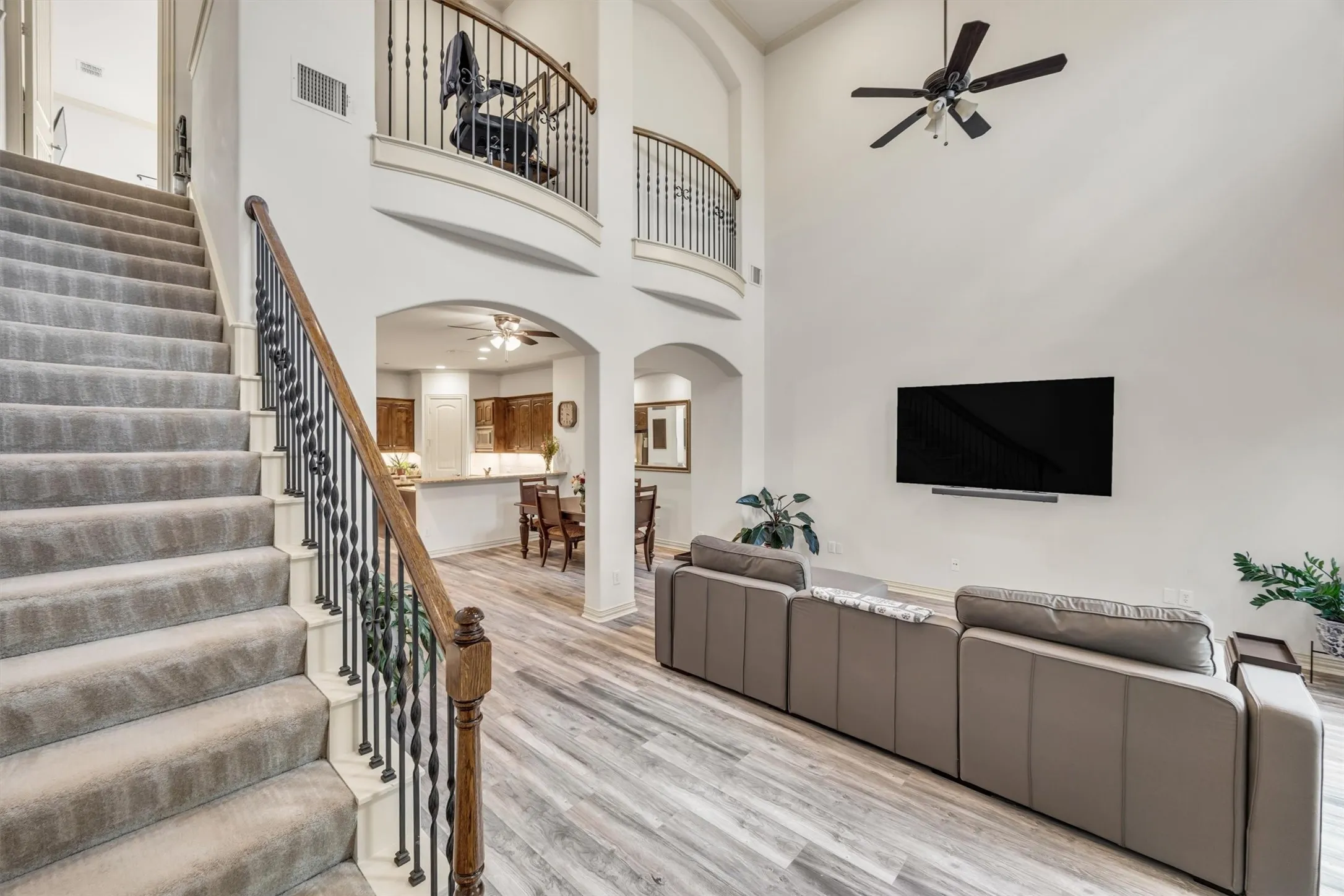Two-story living area with high ceilings, wood-finish flooring, and a carpeted staircase with an ornamental metal baluster