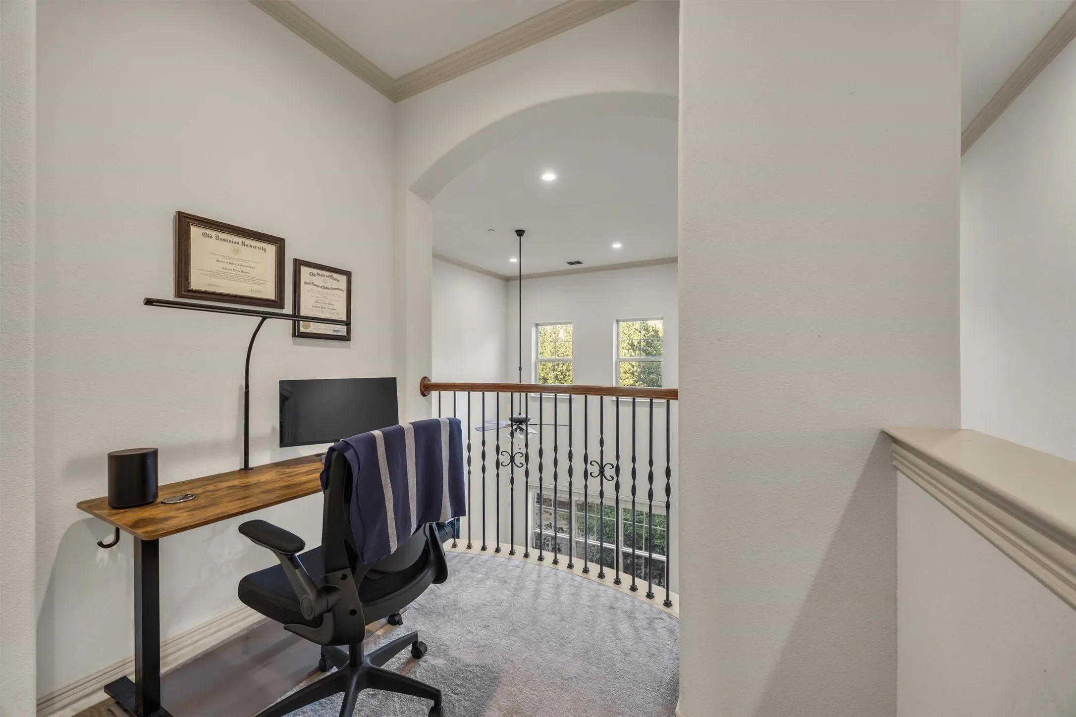 Archway entry to a versatile loft space featuring wrought iron balusters, wood-finish railing, and light-toned carpeting