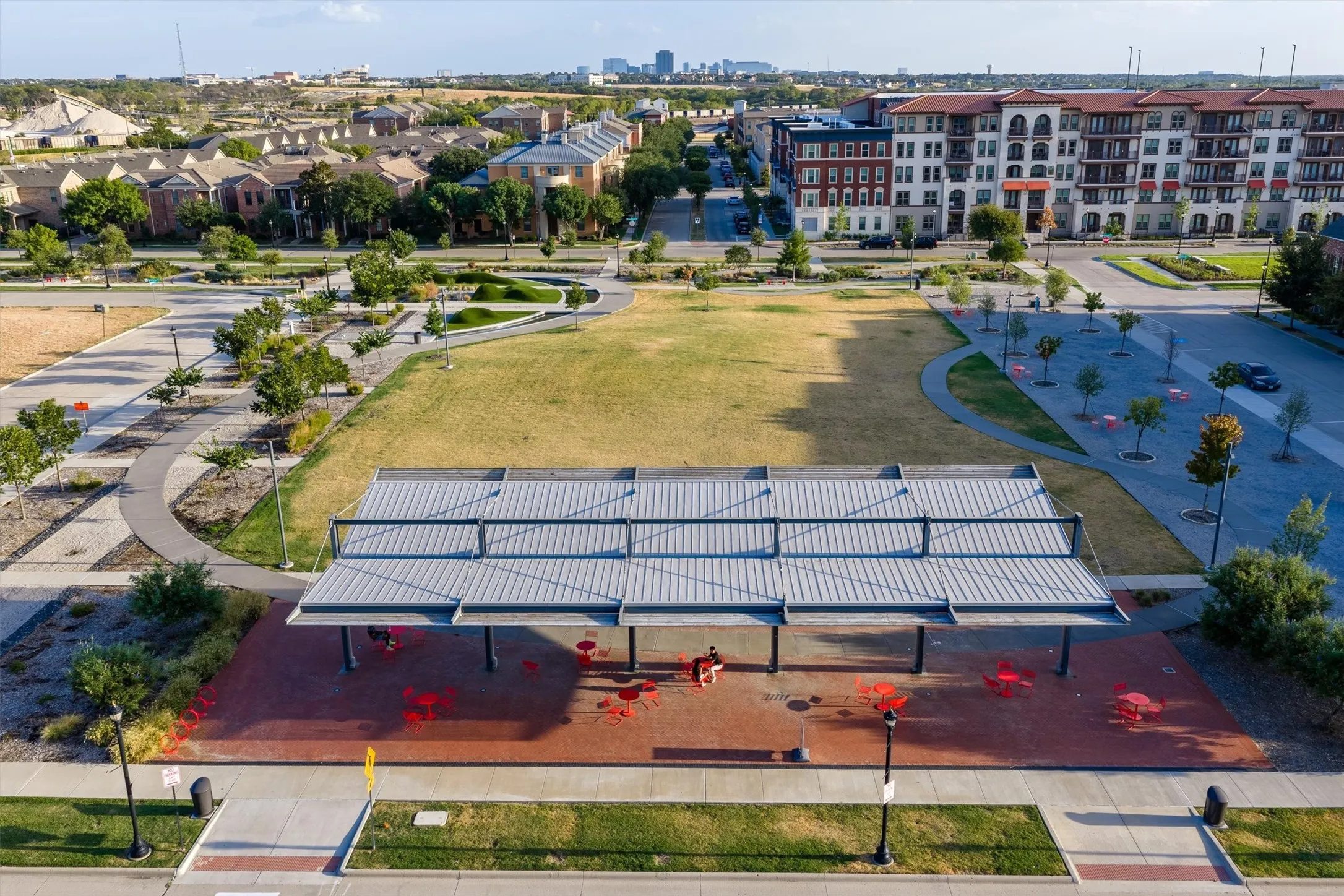 Expansive park area with a large open lawn, paved walking paths, a contemporary open-air pavilion, and structured landscaping featuring trees and shrubbery