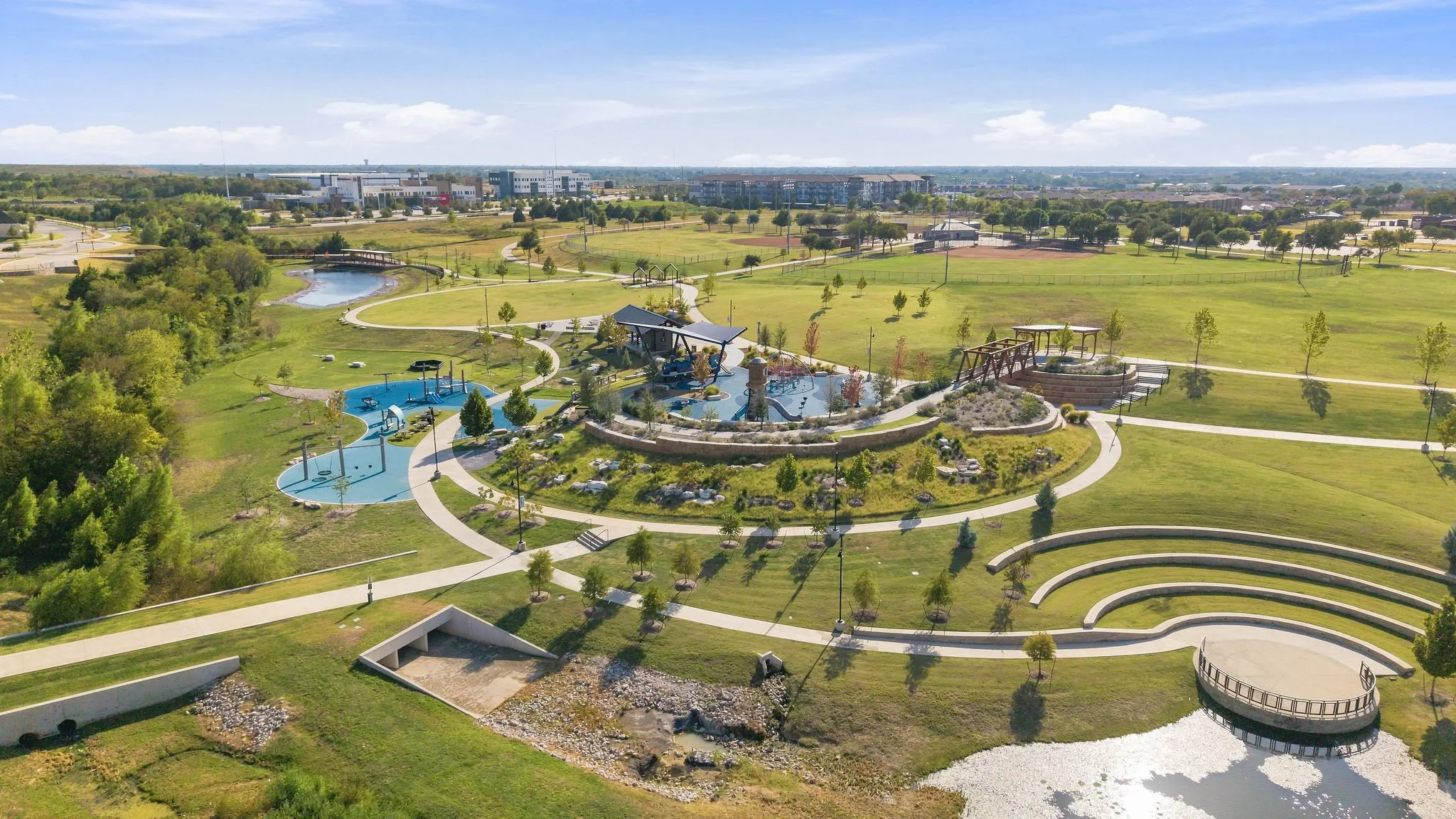 Expansive park setting featuring a tiered amphitheater, a water feature with a stage, extensive walking paths, and a playground area with multiple structures