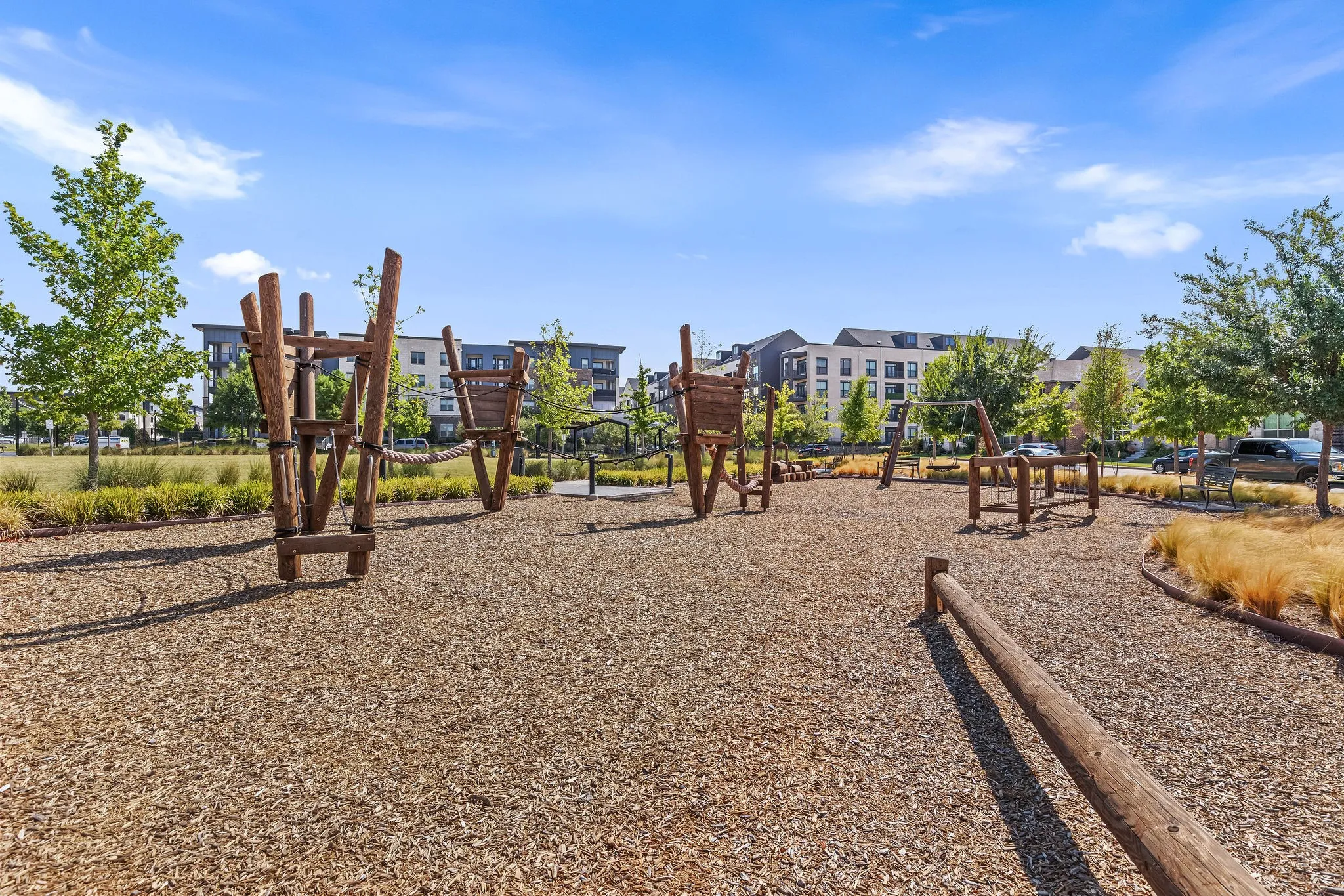 Outdoor recreational area featuring wood chip ground cover, various wood play structures, mature trees, and landscaped borders