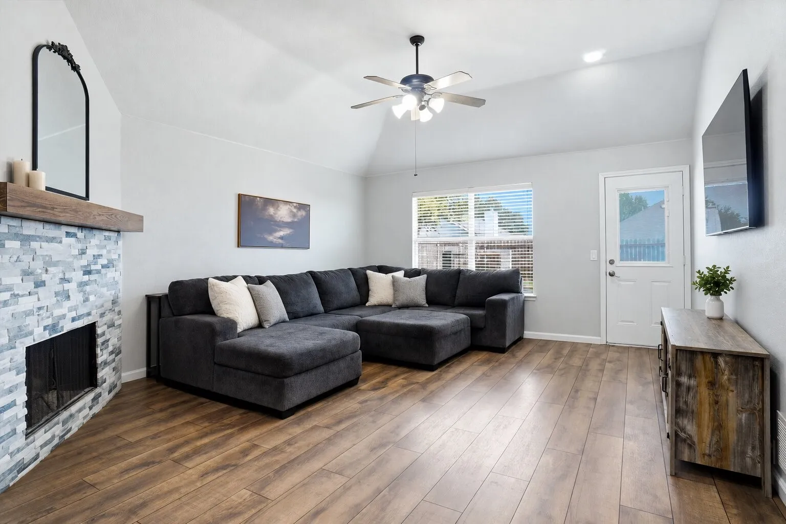 Spacious living room featuring a vaulted ceiling, a floor-to-ceiling stone fireplace, abundant natural light, and direct backyard access. Photos have been AI enhanced for presentation; no material property features were altered.