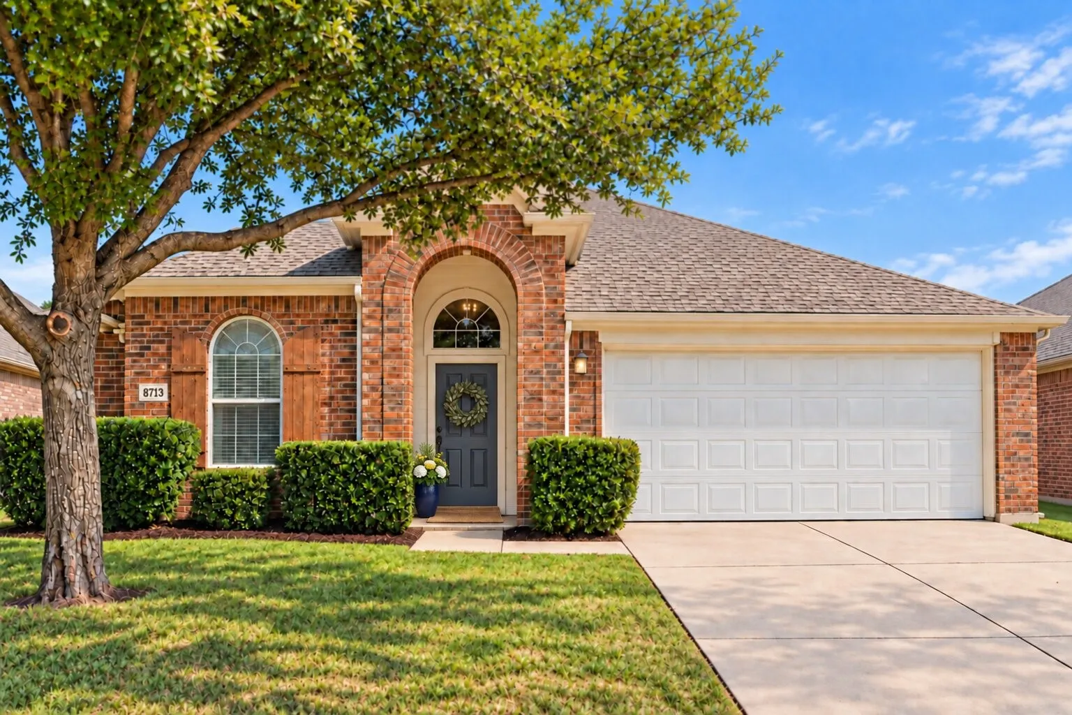 Inviting single-story brick home with mature shade trees, attractive landscaping, an arched entry, and a two-car garage. Photos have been AI enhanced for presentation; no material property features were altered.