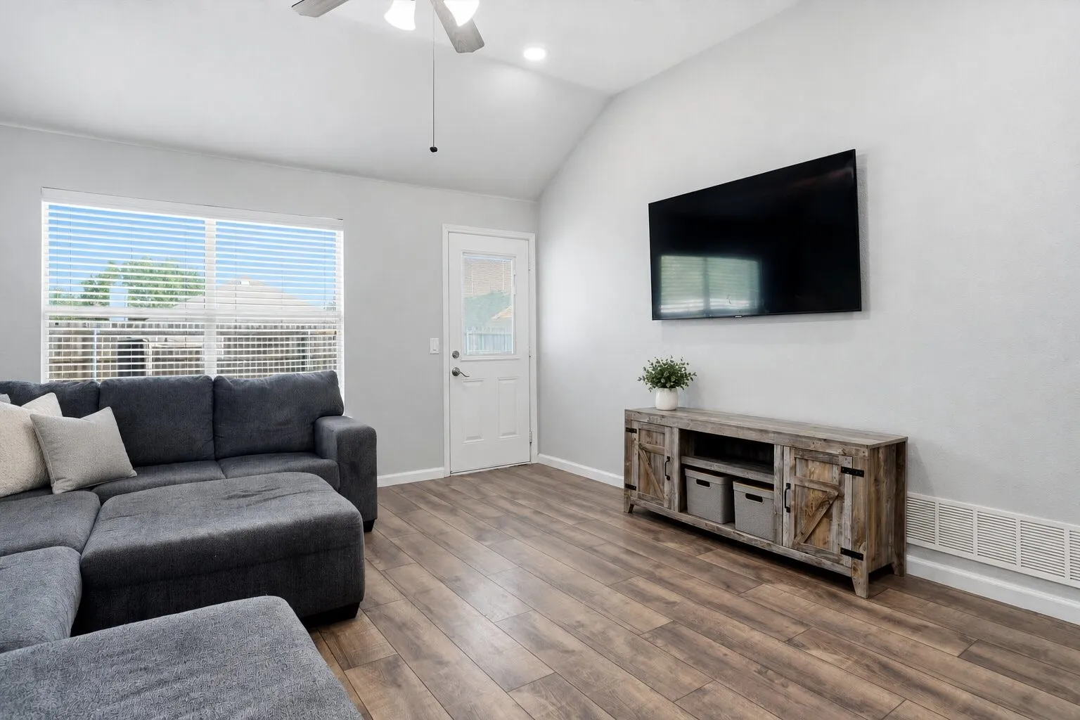 Vaulted living room with abundant natural light, wood-look flooring, a wall-mounted TV, and convenient access to the backyard. Photos have been AI enhanced for presentation; no material property features were altered.