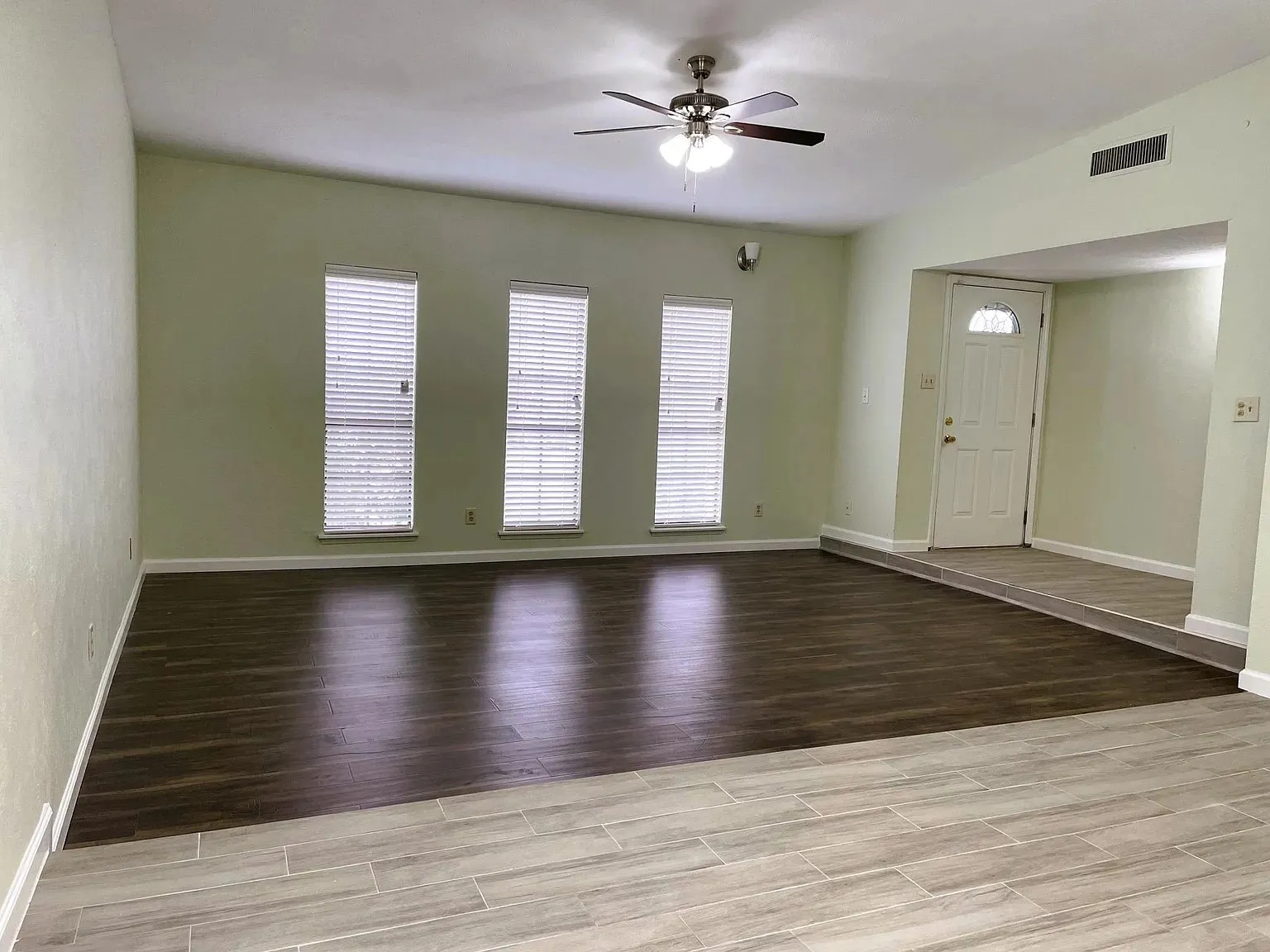 Vaulted ceiling, ceiling fan with integrated lighting, wood-finish flooring, three vertical windows with blinds, and white entry door with upper glass panel