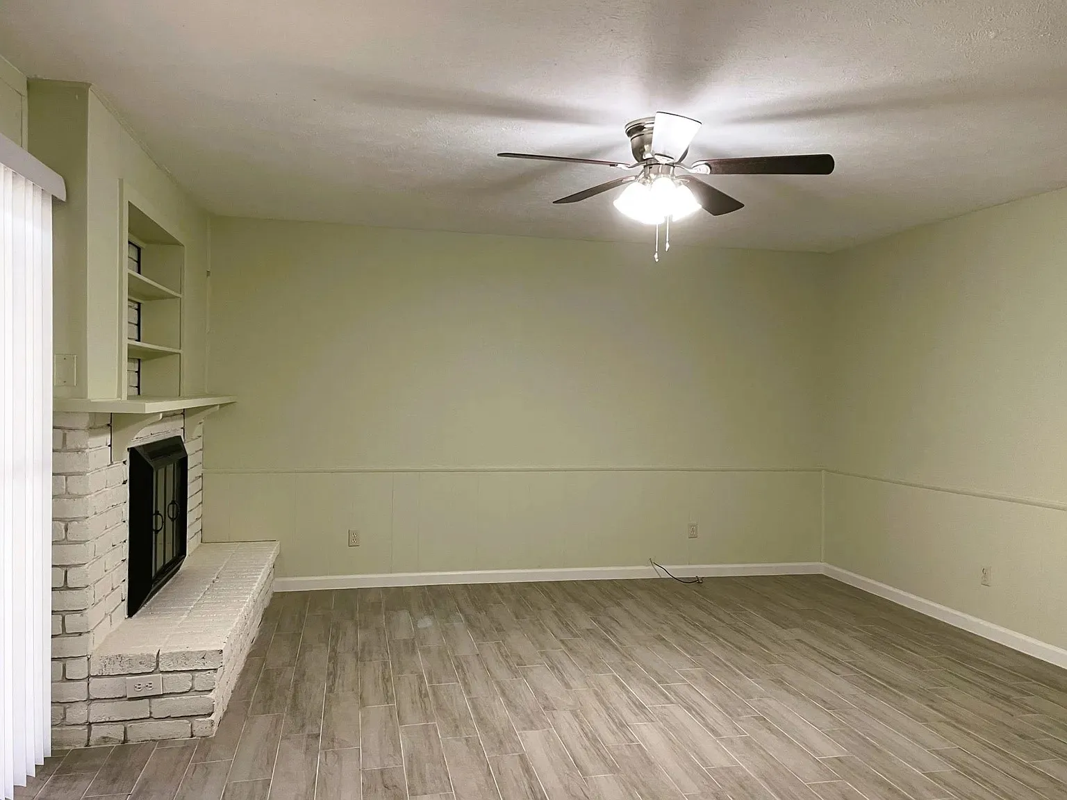 Wood-finish flooring, white brick fireplace with hearth, built-in shelving, and ceiling fan